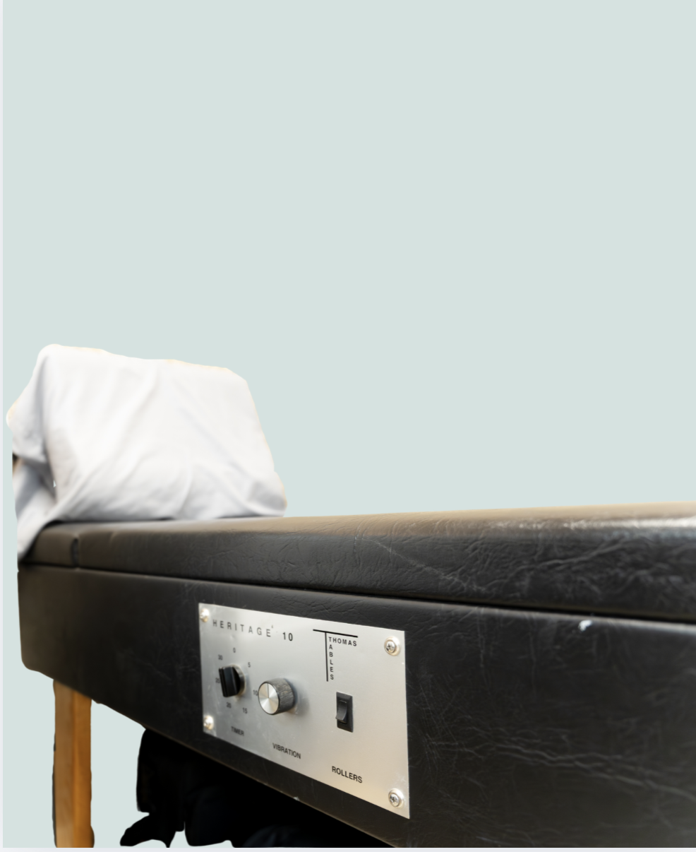 A medical examination table with a white pillow on the left side and control panel on the right side, against a light blue wall.