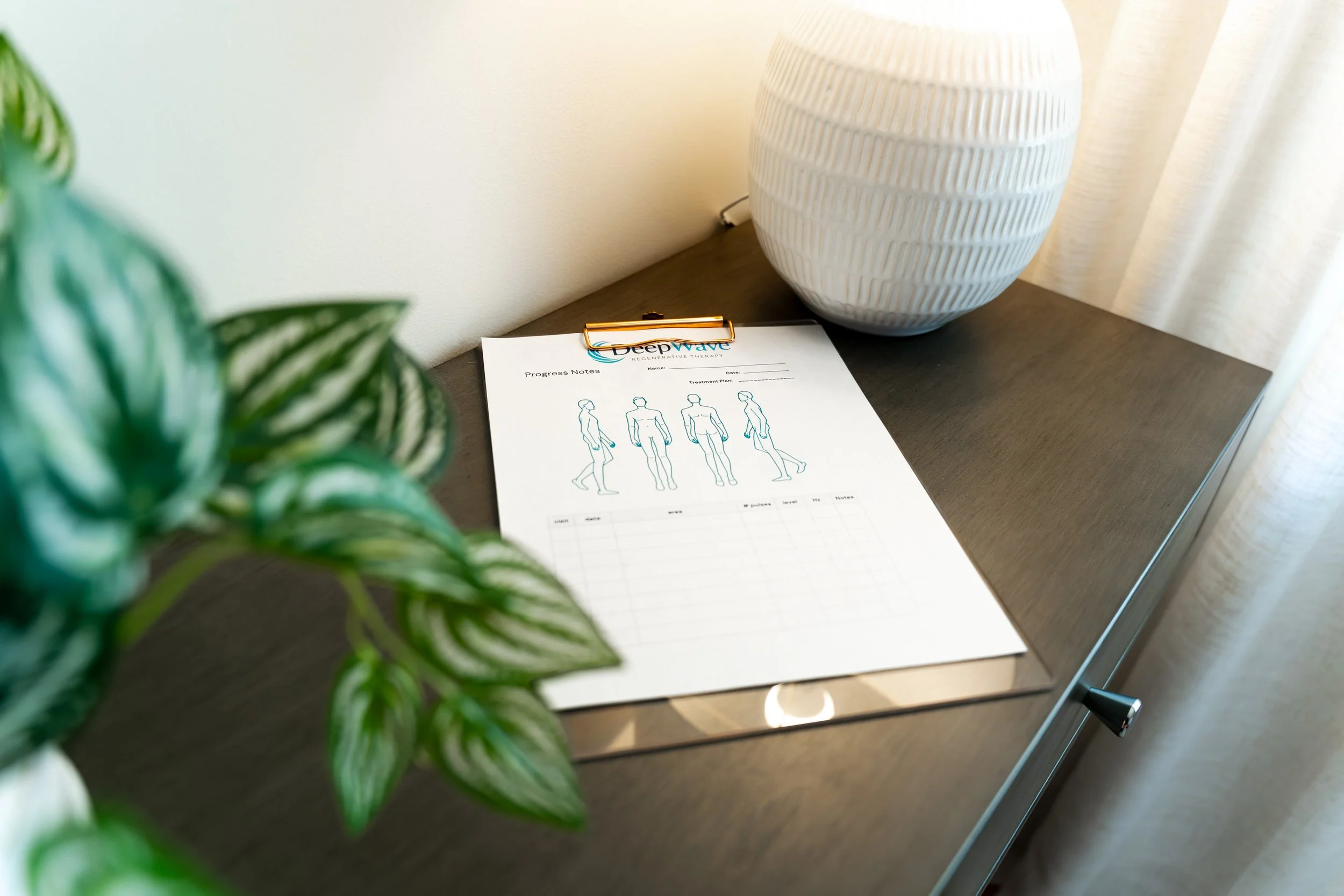 A desk with a potted plant in the foreground, a white vase on the right, a wall in the background, and a sheet of paper with diagrams and notes on it, attached to a clipboard.