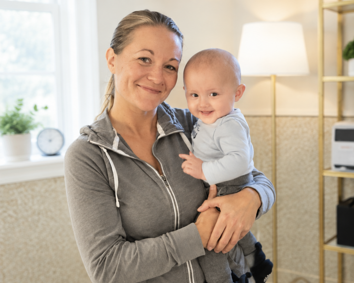 Smiling woman holding a happy baby in a bright room with a window and yellow shelves in the background.
