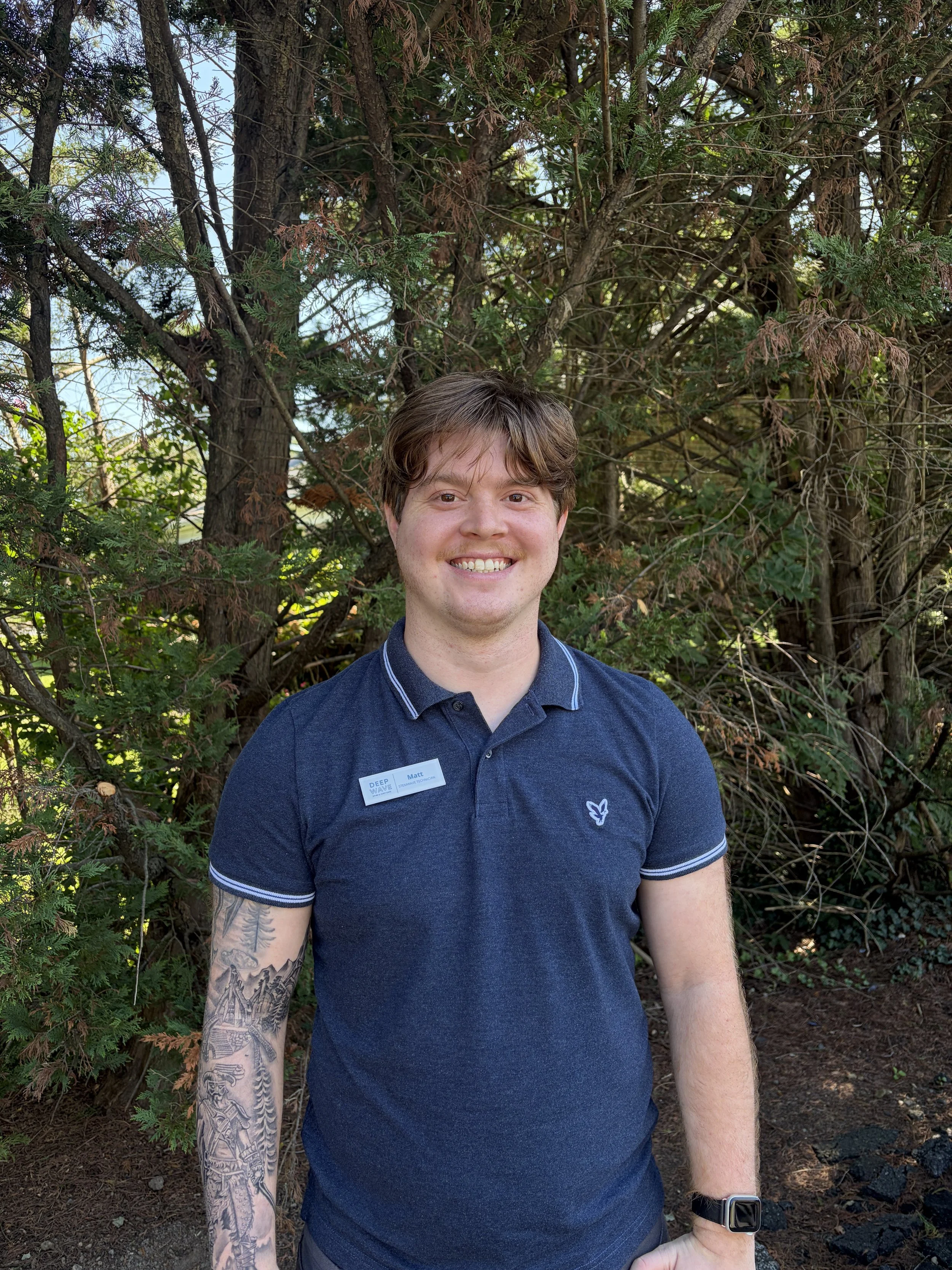 A young man with brown hair and tattoos on his left arm, smiling, wearing a dark blue polo shirt and a watch, standing outdoors in front of a background of trees and greenery.