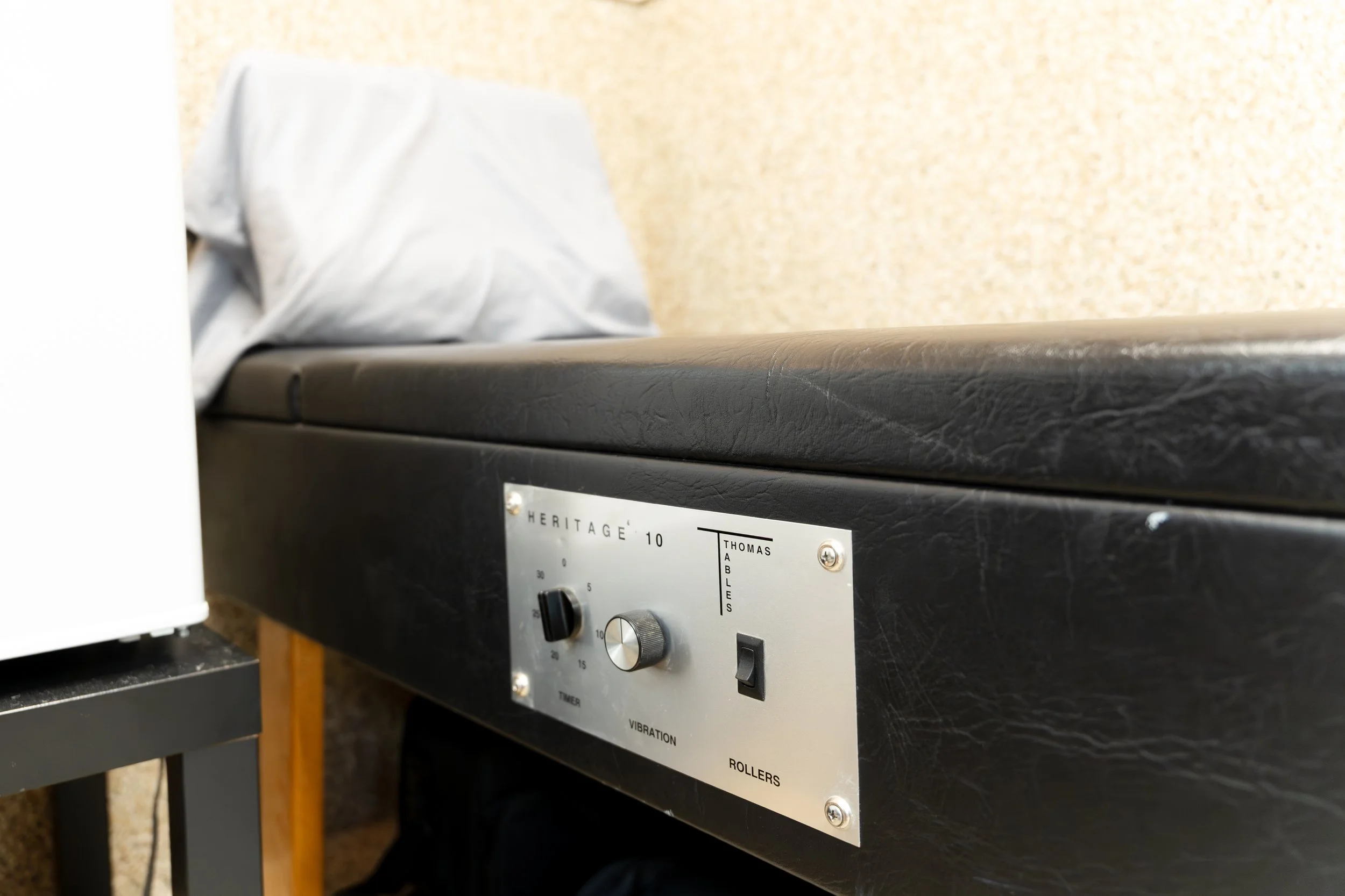 A black machine with a metal control panel featuring black knobs, a switch, and a label reading 'HERITAGE 10'. The machine is placed on a gray table with a yellow wire hanging from it, and part of a white object is visible on the left side. The background includes a beige textured wall.
