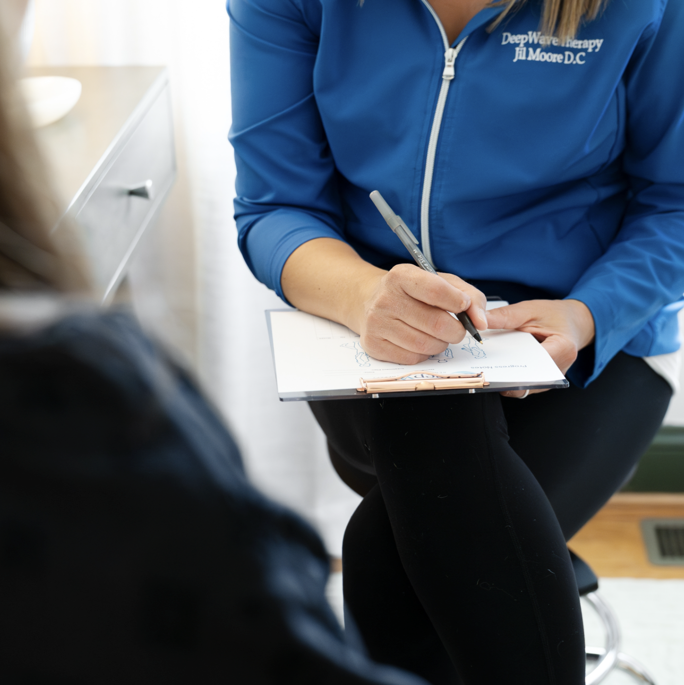 A woman in a blue jacket is sitting and writing on a clipboard with a pen. The jacket has white text that reads 'Deep Wave Therapy Jill Moore D.C.'