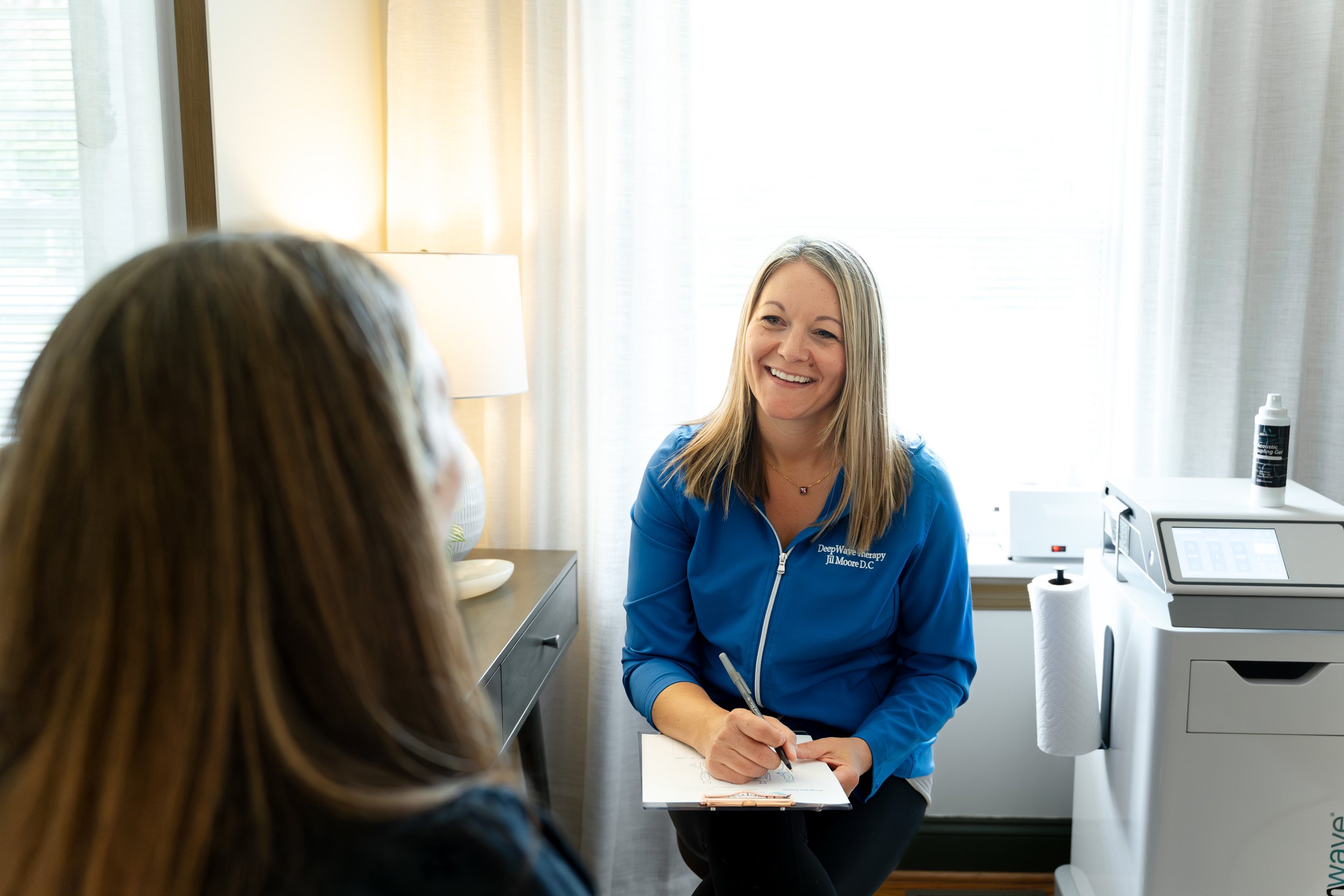 A smiling female healthcare professional in blue scrubs sitting in a consultation with a patient in a bright room, with medical equipment visible in the background.