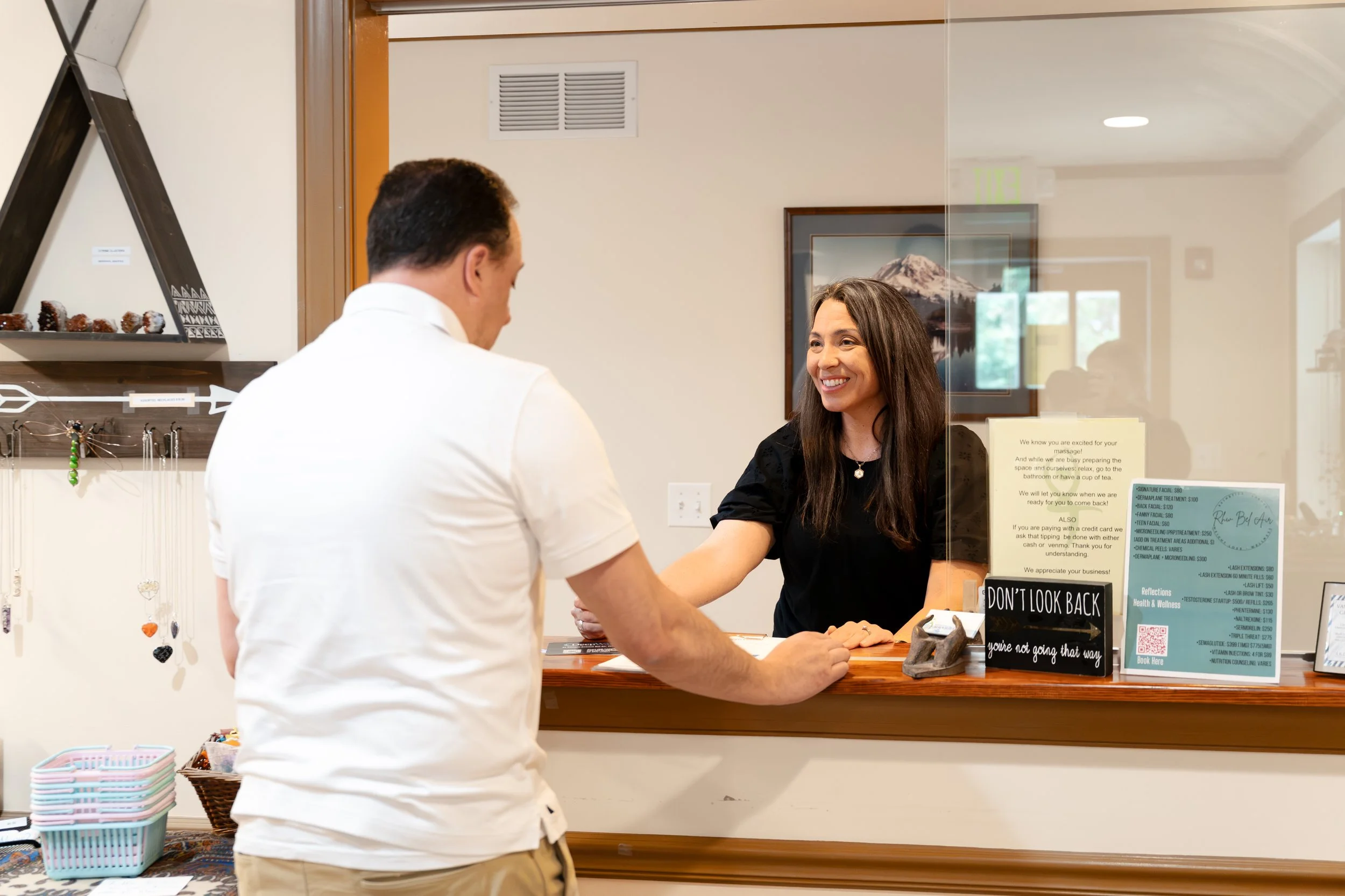 A woman at a reception desk smiling and shaking hands with a man. The reception area has a glass divider, a framed mountain photograph, and various informational signs.