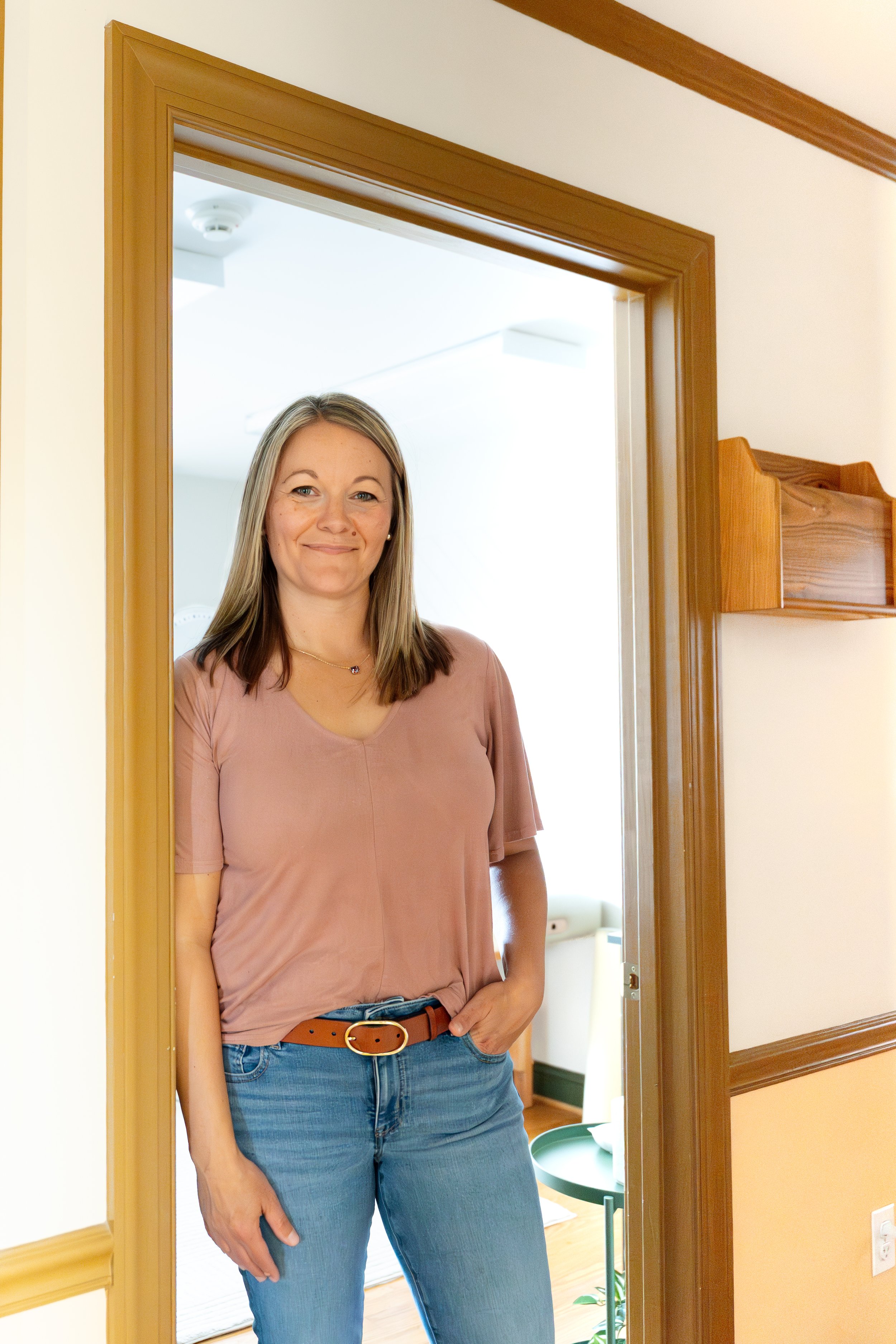 A woman smiling and standing in a doorway in a home interior. She has shoulder-length blonde hair, is wearing a pinkish-beige top and blue jeans with a brown belt, and has one hand in her pocket.