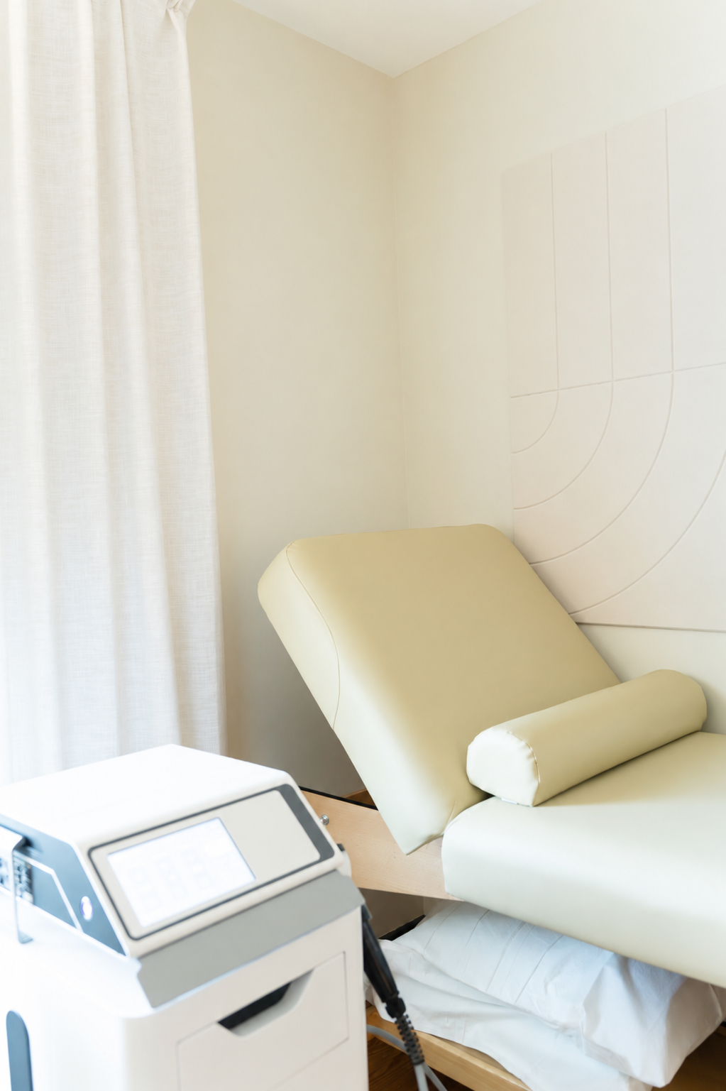 A medical examination room with a beige reclining examination chair, a white curtain, and medical equipment near the chair.