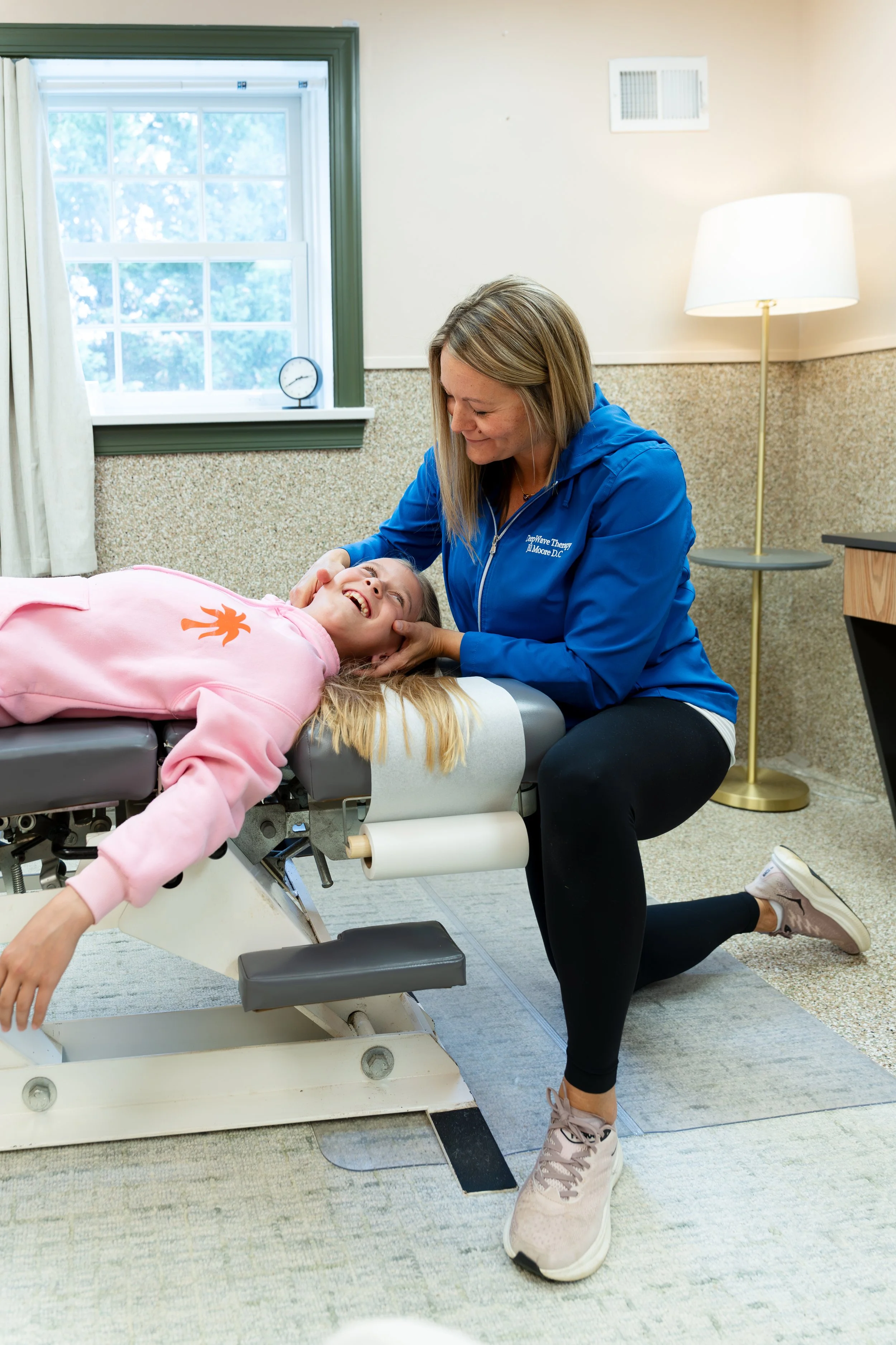 A woman in a blue jacket kneels beside a smiling young girl lying on a physical therapy table, holding her head and shoulder while performing a therapy session in a room with a window, curtains, floor lamp, and patterned walls.