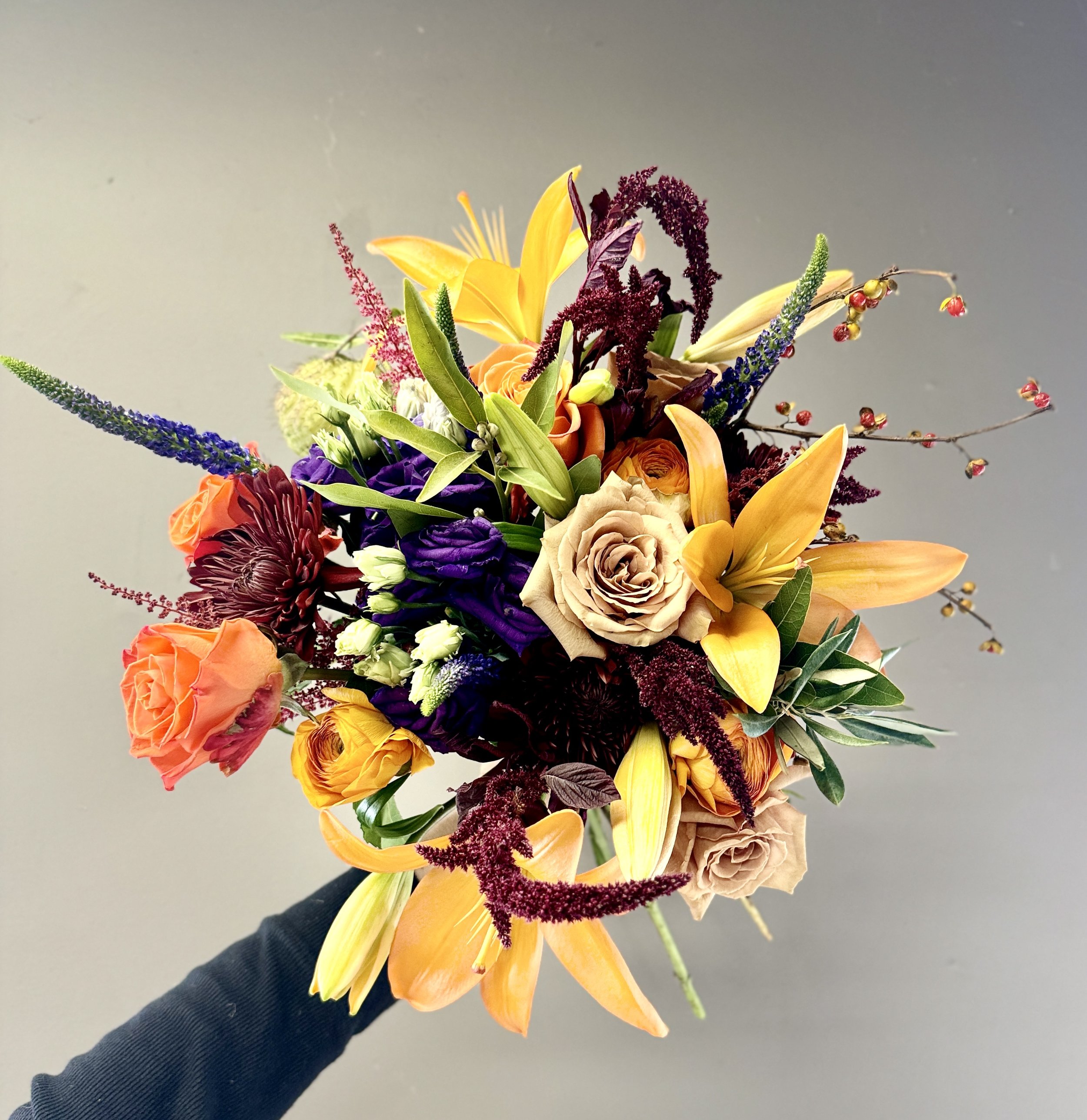 Hand holding a colorful bouquet of various flowers including yellow lilies, orange roses, purple statice, and red berries, against a gray background.