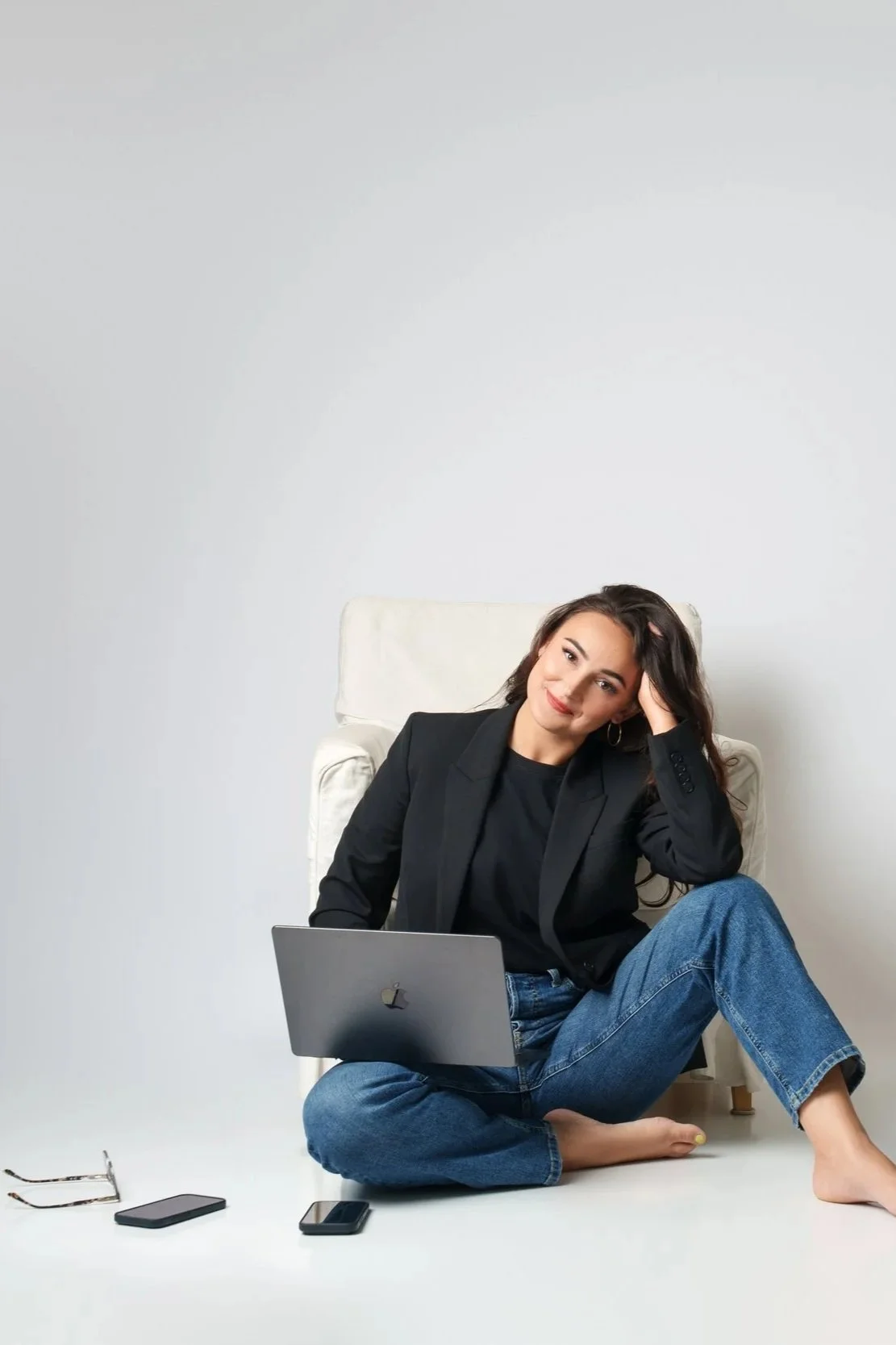 Young woman sitting on the floor with a laptop on her lap, smiling and resting her head on her hand, with smartphones and glasses on the floor around her, against a plain white background.