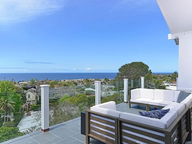 View from a balcony showing the ocean, houses, trees, and a clear blue sky with a few clouds, with a white outdoor couch on the balcony.