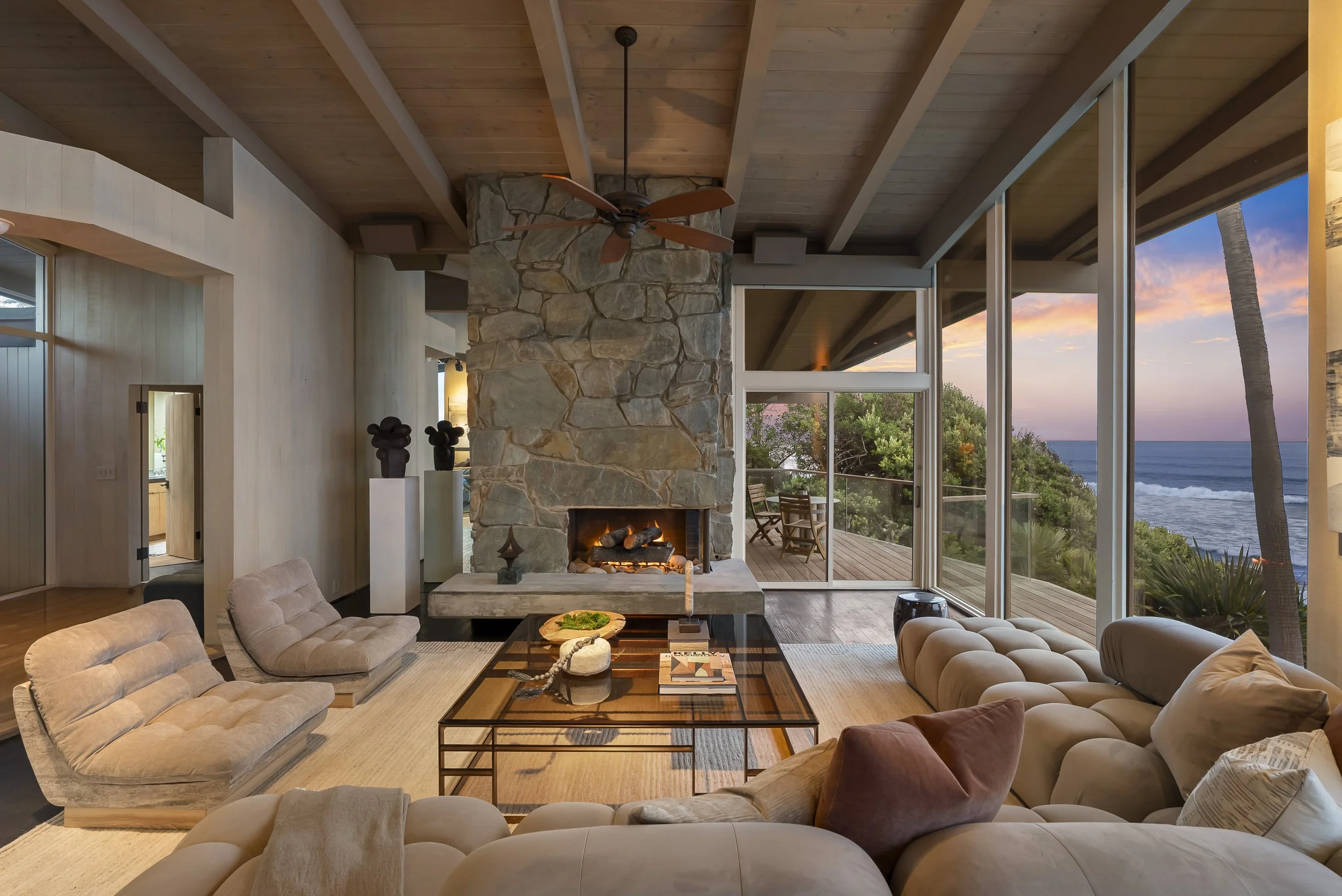 Living room with beige sofas, a glass coffee table, stone fireplace, large windows showing a view of the ocean, palm trees, and a sunset sky.