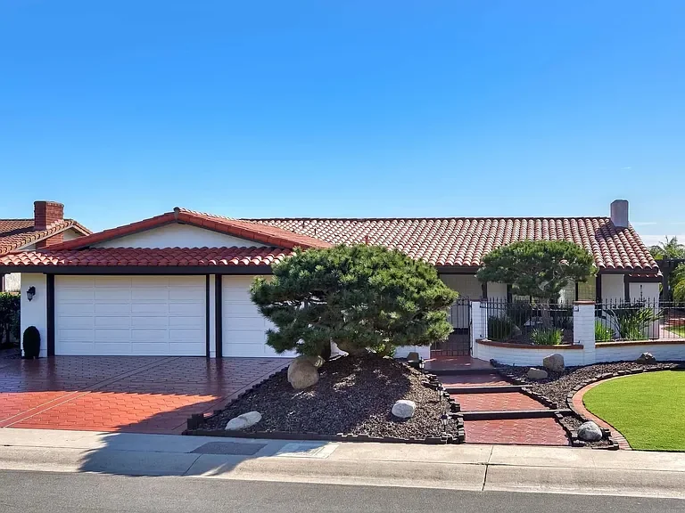 Single-story house with a red-tiled roof, white walls, garage door, and a front yard featuring a neatly trimmed tree, rocks, and a manicured lawn under a clear blue sky.