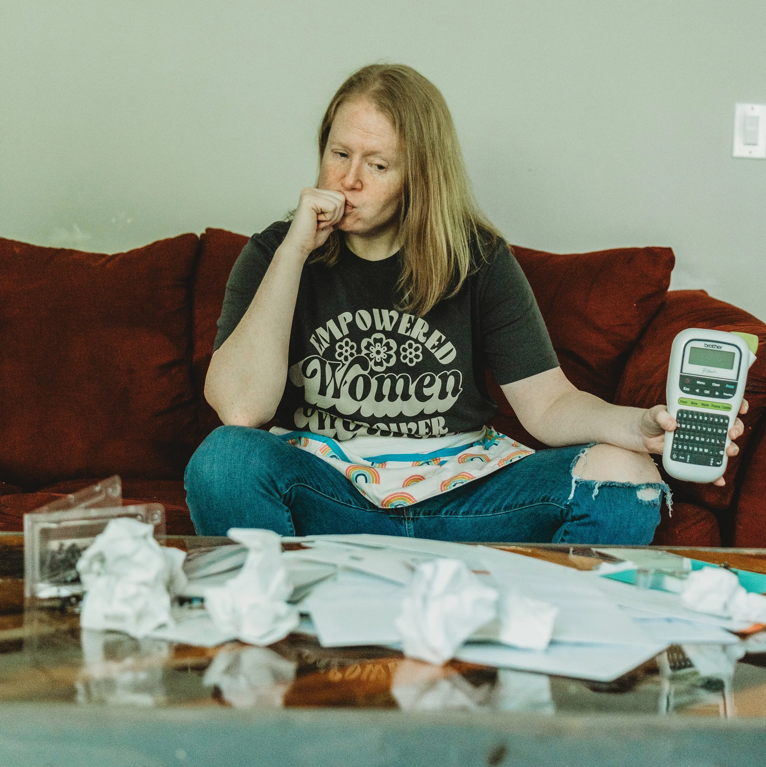A woman with ginger hair sitting on a red couch, wearing a black T-shirt with white text, holding a label maker, looking thoughtfully at a lot of crumpled paper on a table in the foreground.