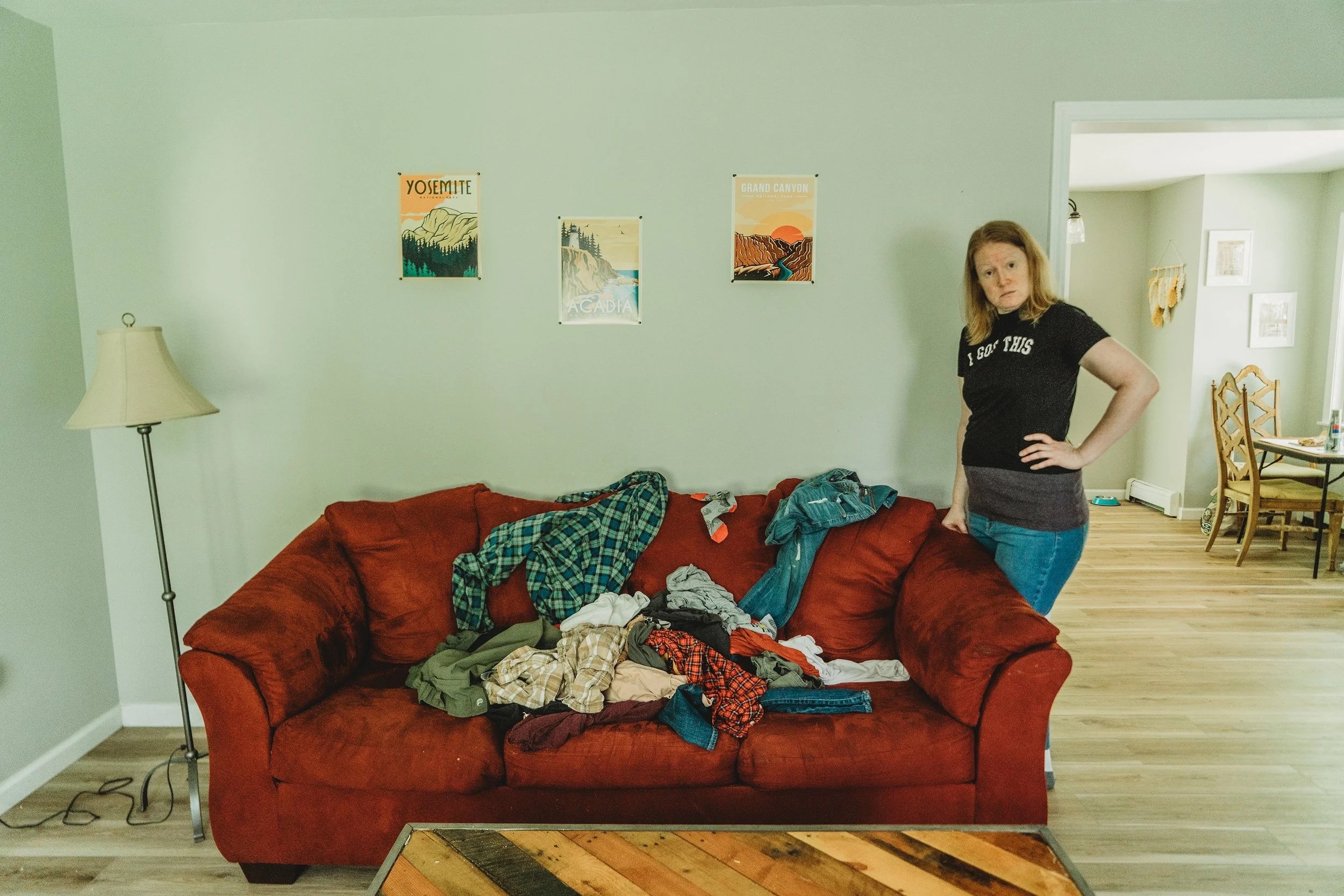 A iving room with a standing lamp and a red sofa. The sofa is covered in messy clothes, and a standing lamp. Woman with ginger hair in a black t-shirt and jeans is standing next to the sofa.