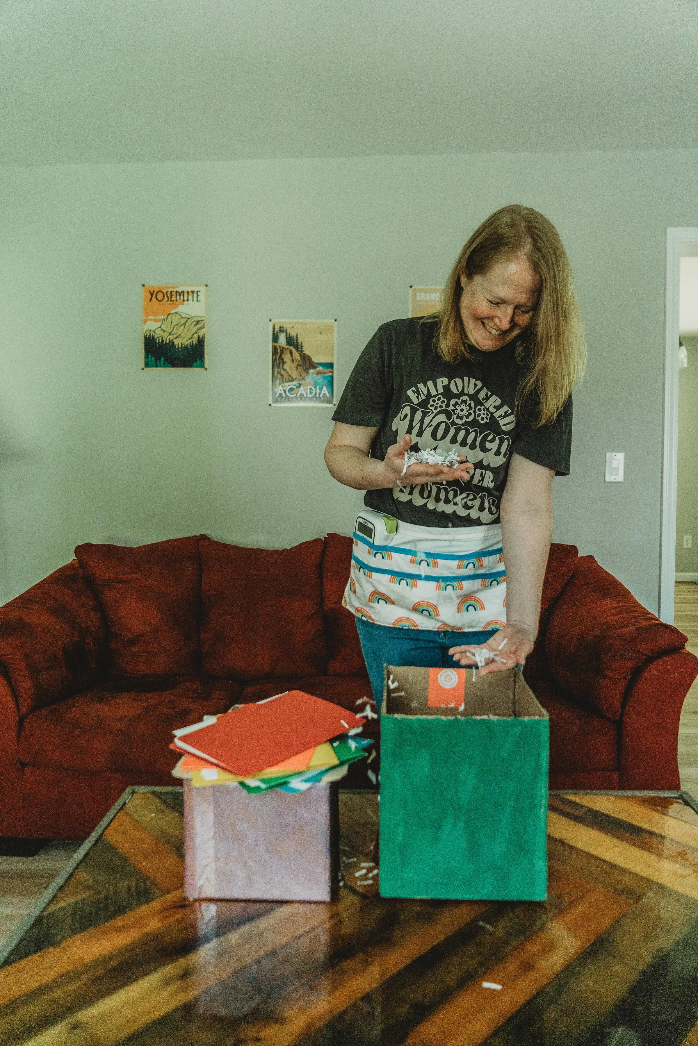 A woman with ginger hair wearing a black shirt with white text, a colorful rainbow apron, is sifting shredded paper next to a stack of tidy, colorful file folders in a living room with a red couch.