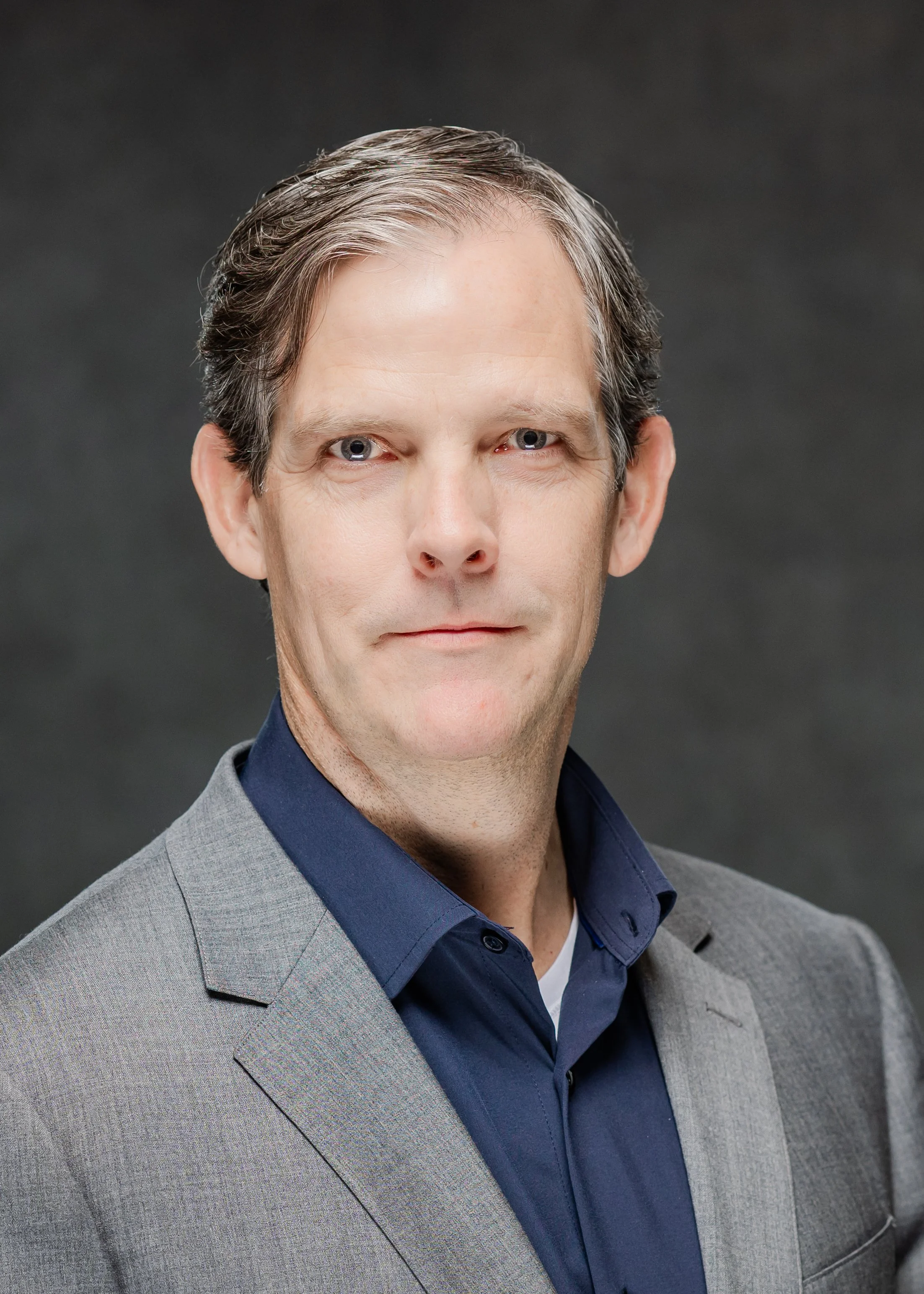 A headshot of a man in a gray suit and blue shirt against a dark gray background.