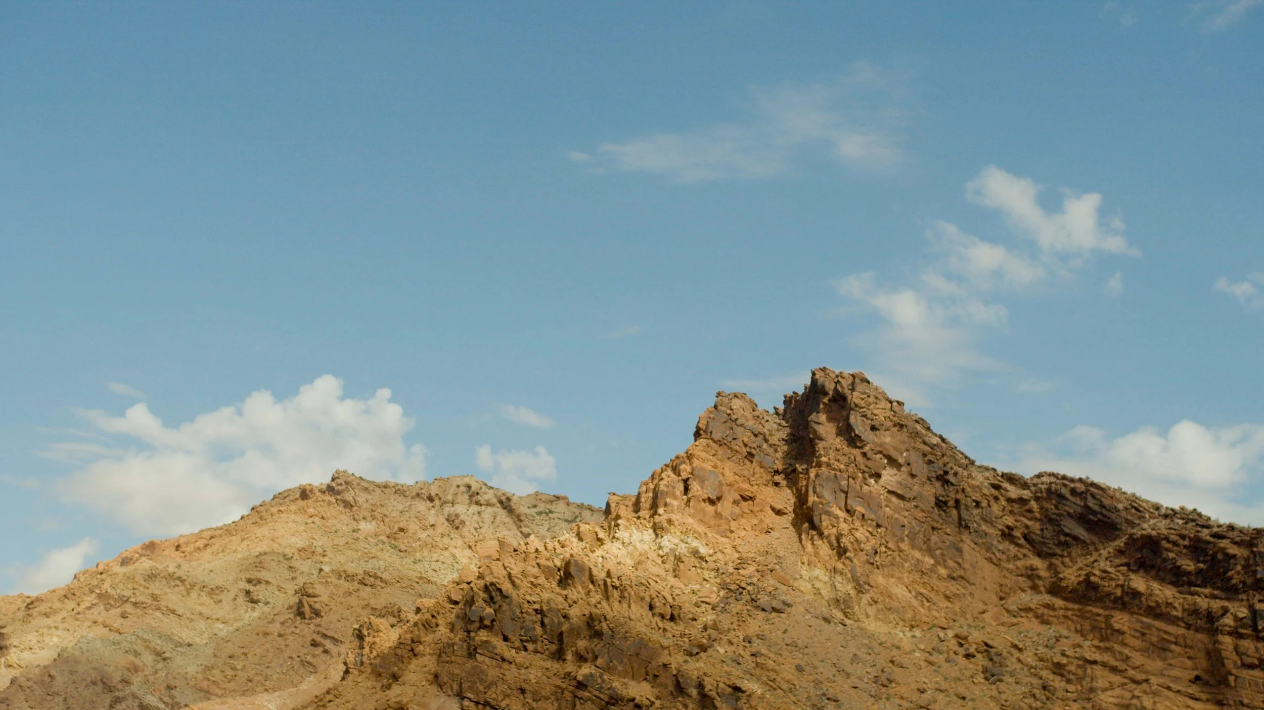 A rugged, rocky desert mountain under a partly cloudy blue sky.