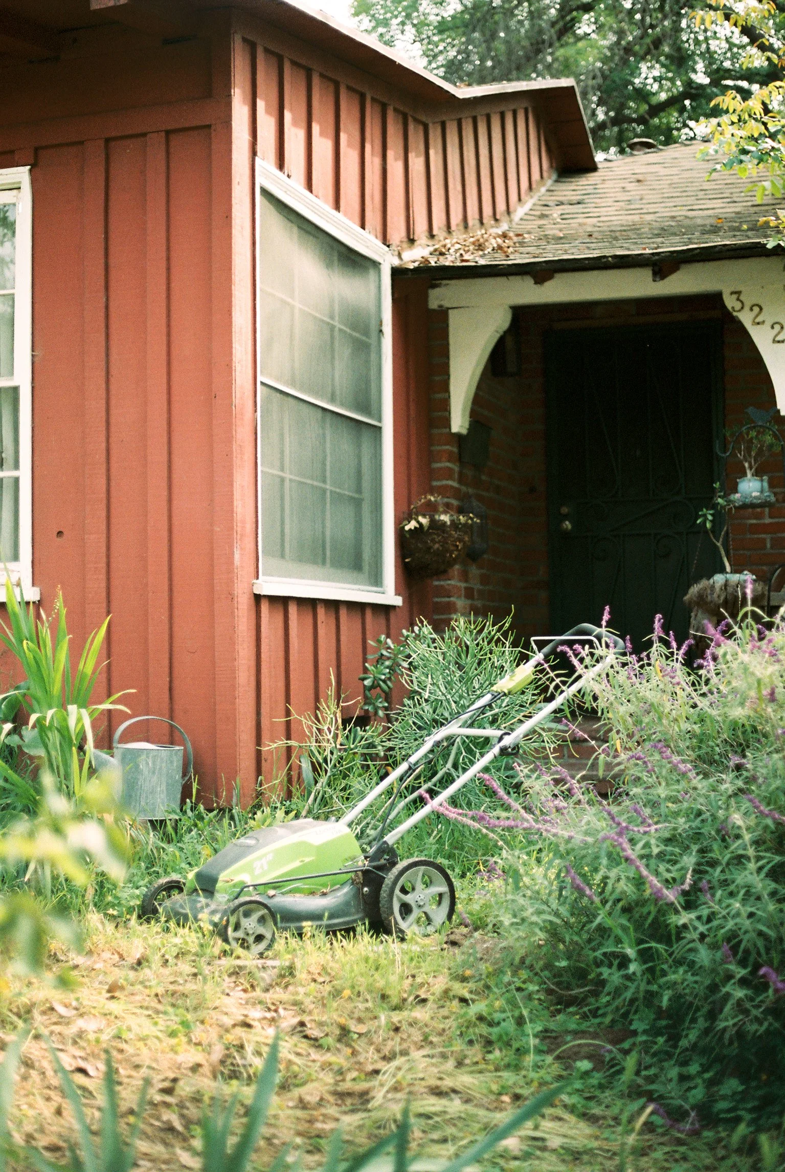 A small green toy lawn mower placed on the grass in a garden with various plants and flowers, in front of a house with a red wooden exterior and a black front door.