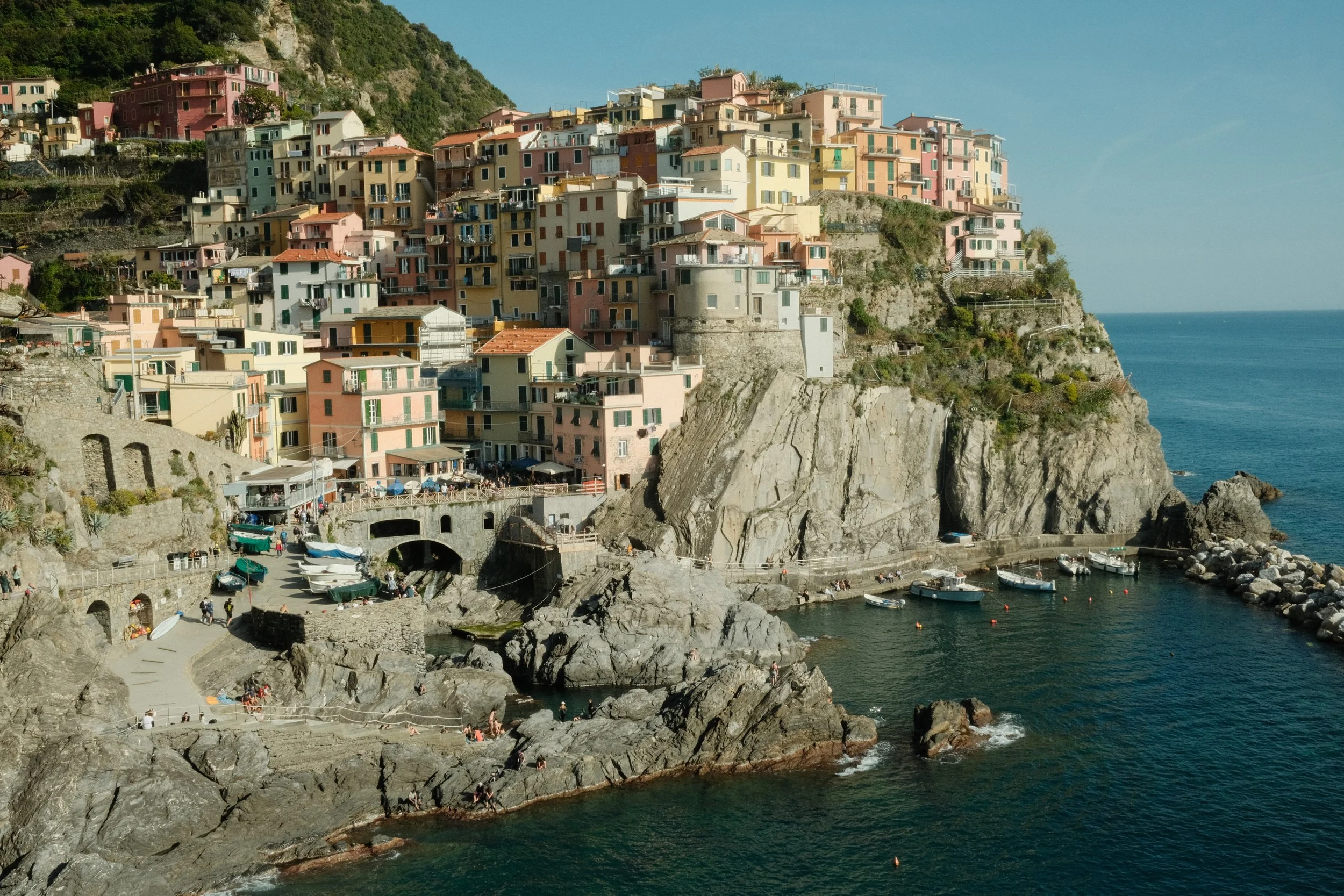 Colorful buildings built on a steep hillside overlooking the Mediterranean Sea with small boats docked at a rocky shoreline and people enjoying the beach.