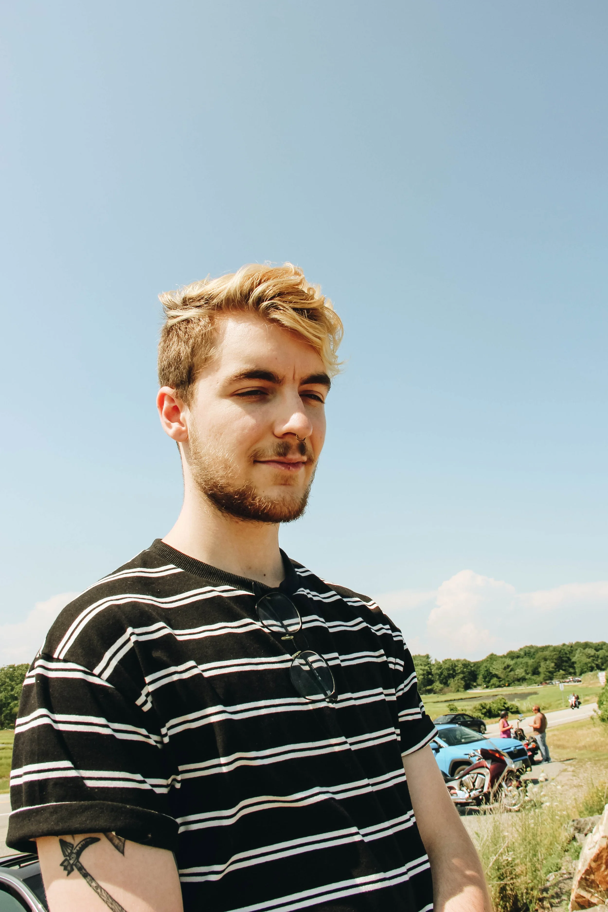 Young man with blonde hair, beard, and tattoos on arm wearing a black and white striped shirt standing outdoors in a grassy area on a sunny day, with cars and people in the background.