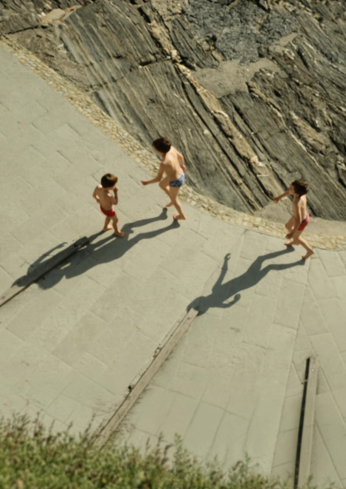 Three children in swimsuits walk along a paved pathway next to a rocky wall, with their long shadows cast on the ground.