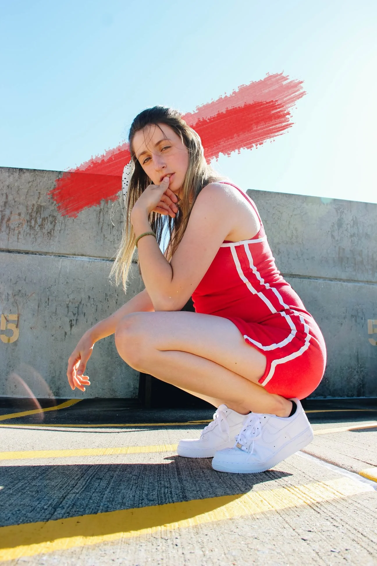 A young woman with long hair wearing a red athletic dress and white sneakers, squatting on a parking lot ground near a concrete wall with colorful markings, with a painted red brush stroke behind her and a clear blue sky above.