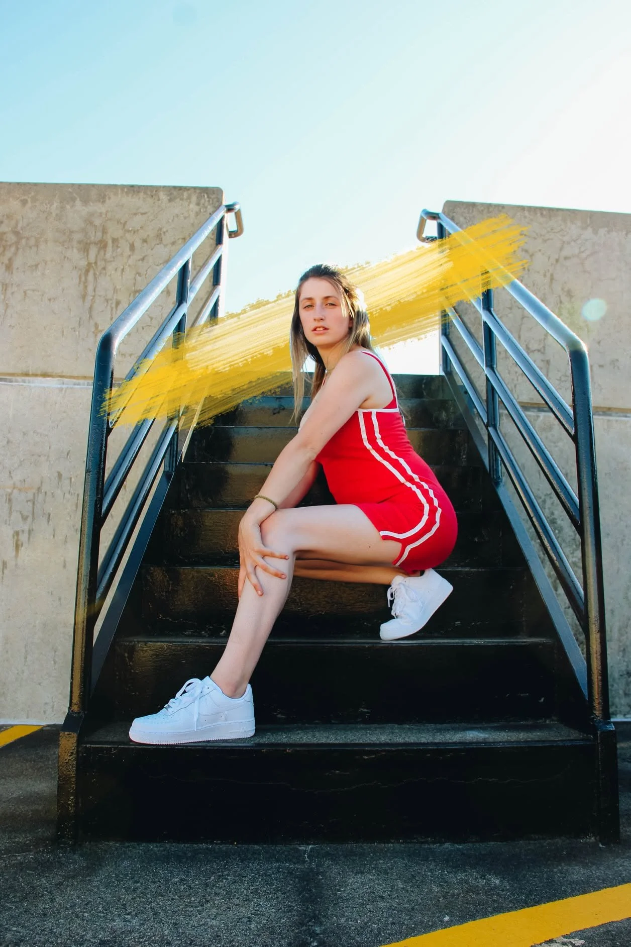 Young woman sitting on outdoor metal staircase with yellow streak across image, wearing red athletic outfit and white sneakers, against a beige wall and blue sky.