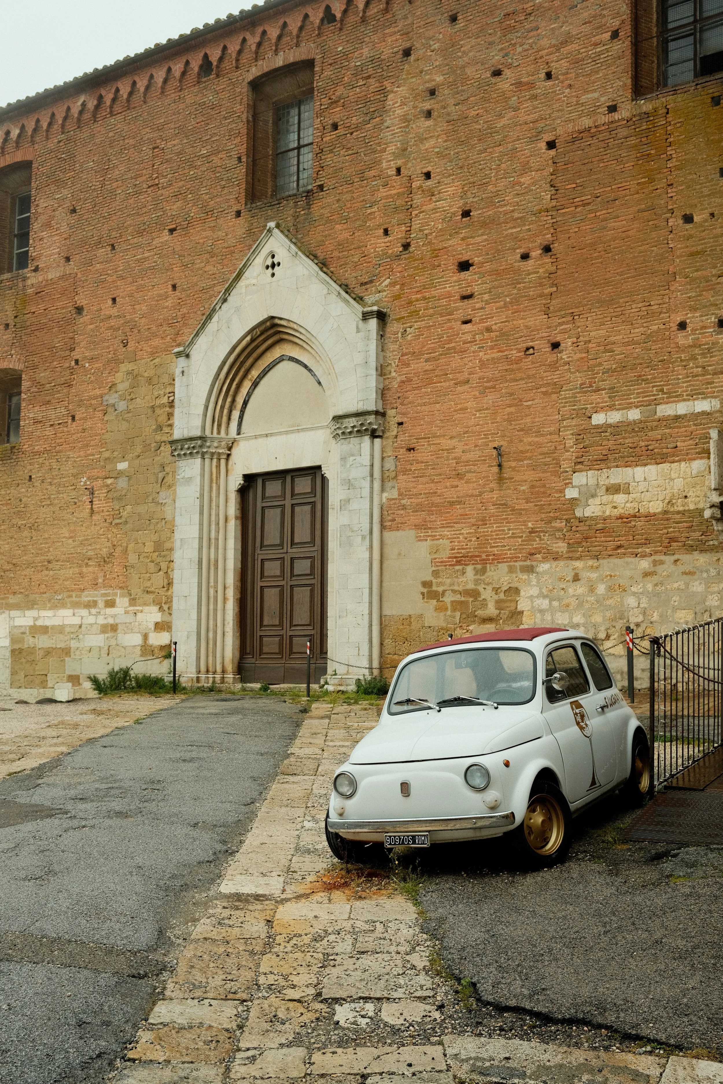 A historic brick church with a large arched wooden door and a white stone frame. A vintage white Fiat 500 car parked in front with a Roman license plate. Cobblestone path leading to the church entrance, with some grass and plants at the edges.