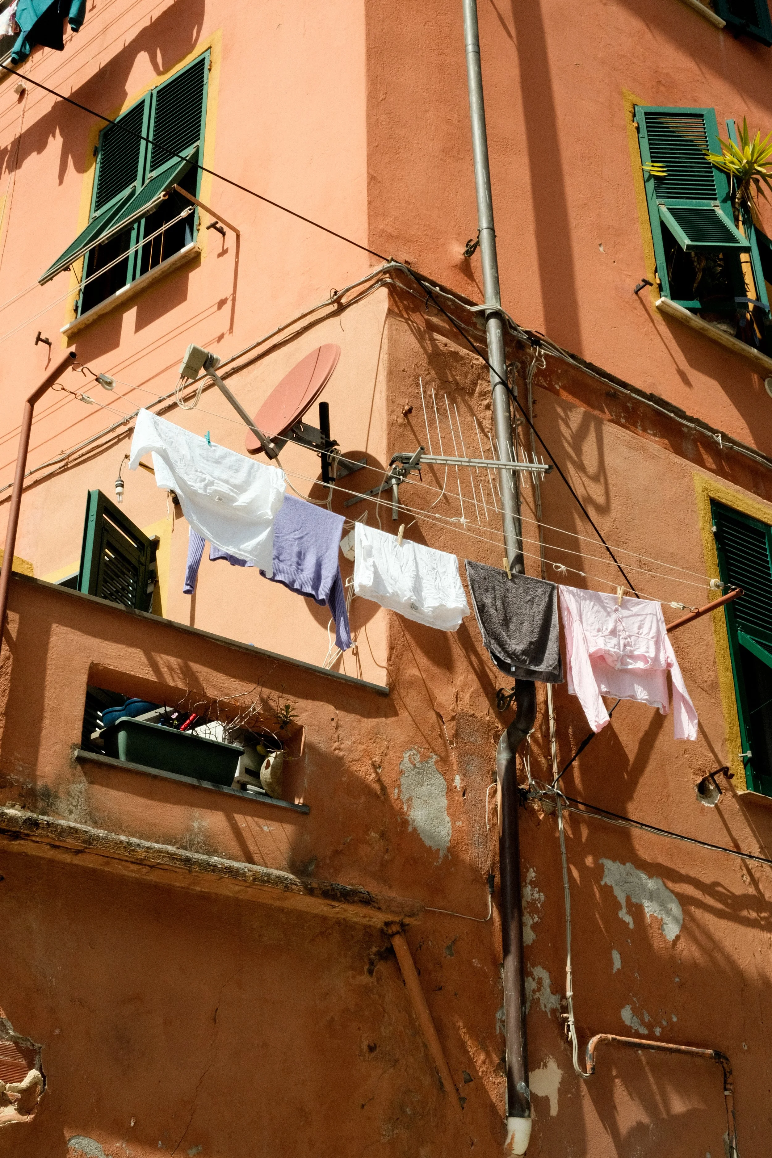 A peach-colored building with green window shutters, laundry hanging on clotheslines, a satellite dish, and a potted plant on the balcony, with visible weathered walls and exposed pipes.