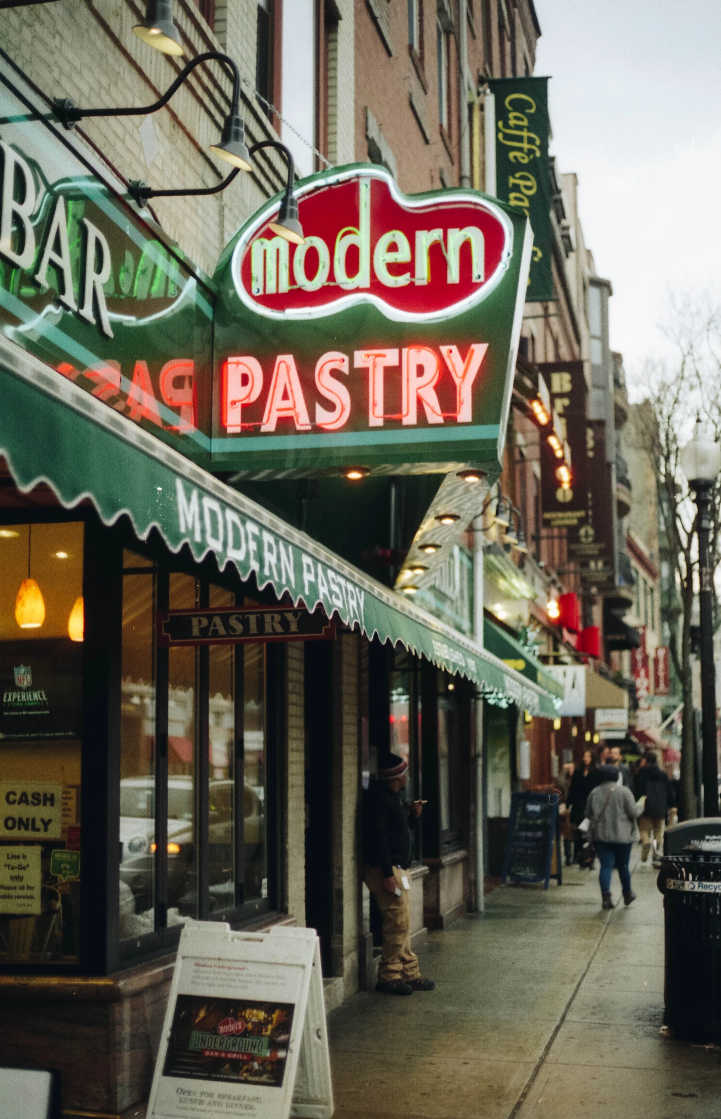 Street view of a storefront with a neon sign that says 'modern' and 'pastry'. There are people walking on the sidewalk and other signs and buildings in the background.