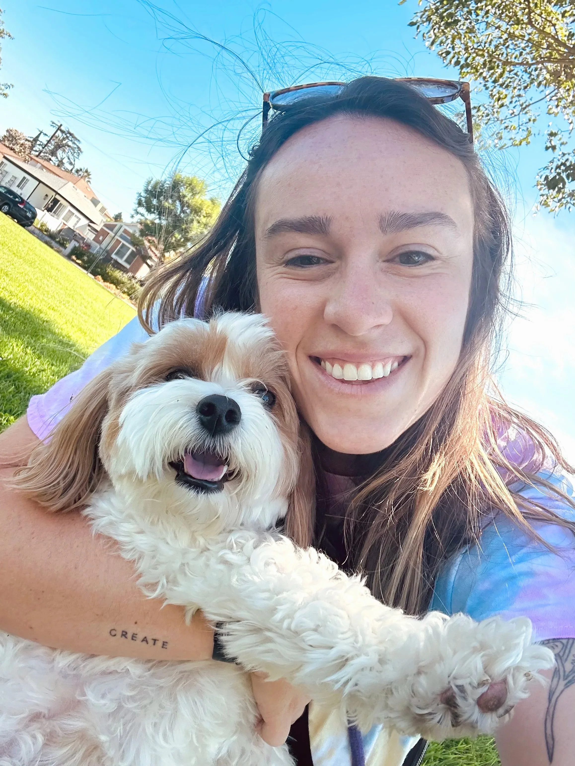 A woman smiling holding a cute fluffy white and brown dog outdoors on a sunny day with blue sky and trees in the background.