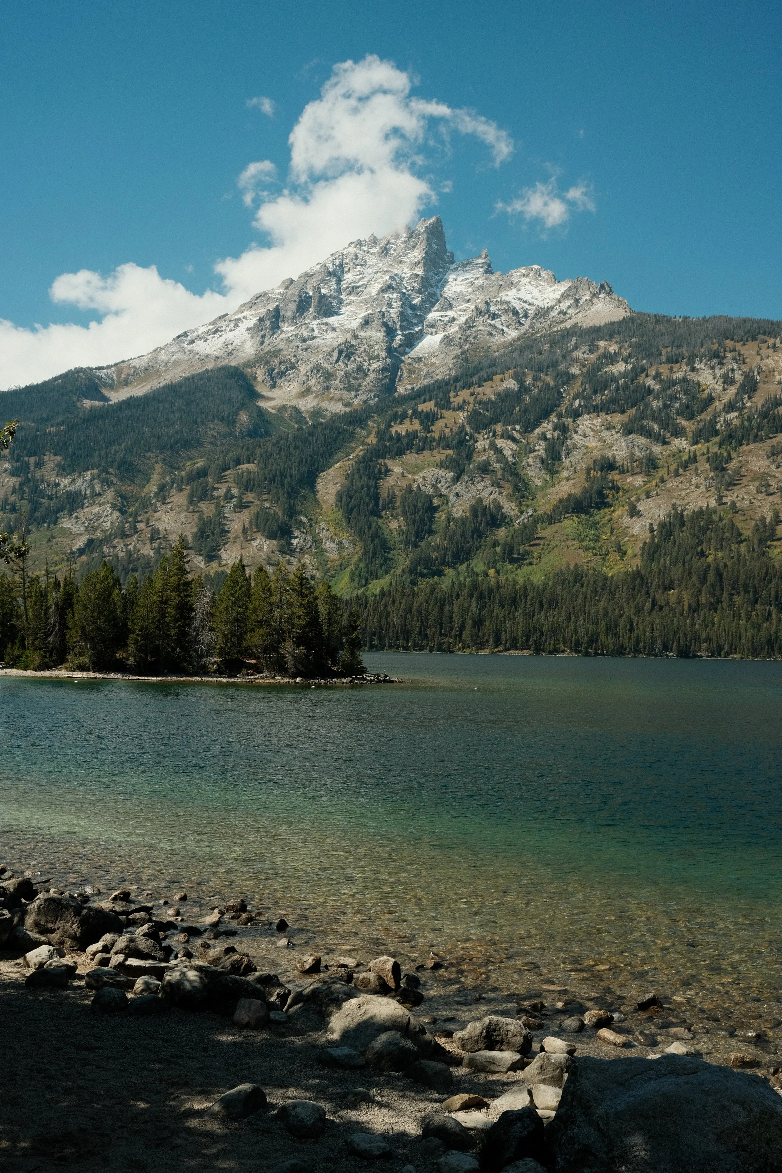 Scenic view of a mountain with rocky peaks, surrounded by forest, next to a calm lake with a rocky shoreline under a blue sky with scattered clouds.