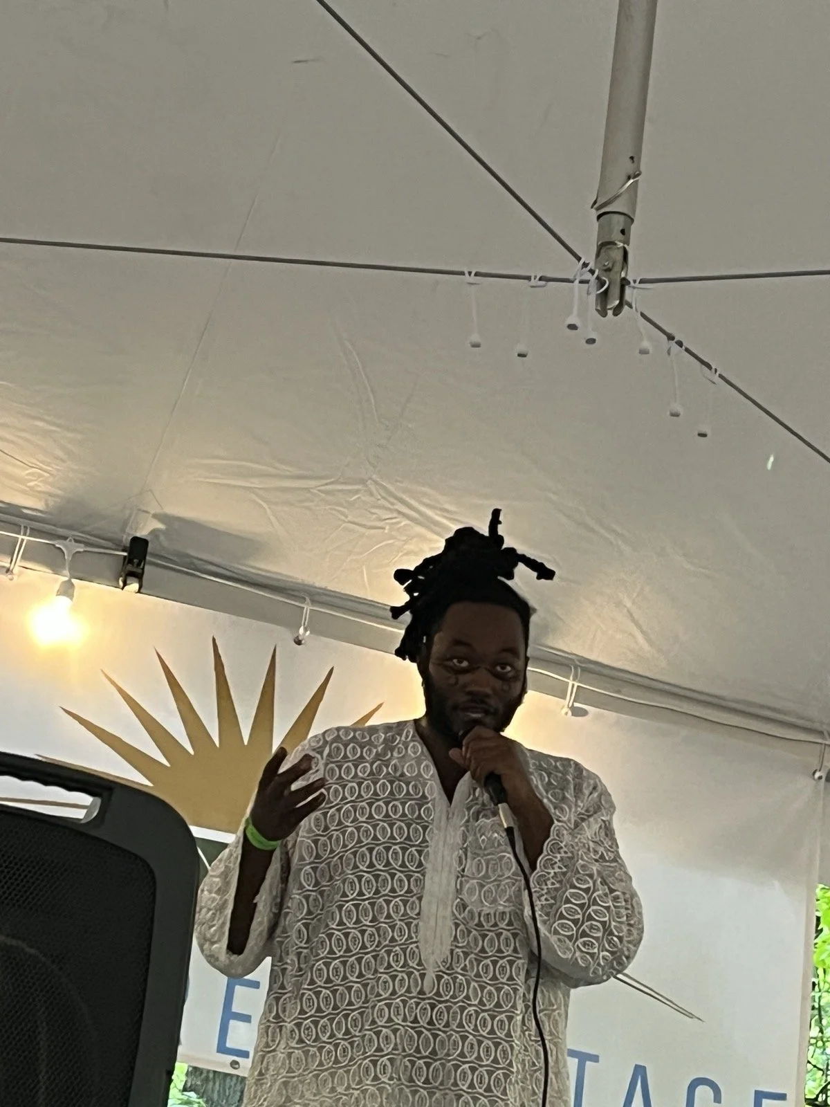 A man with dreadlocks and glasses speaking into a microphone at an indoor event, wearing a white patterned shirt.