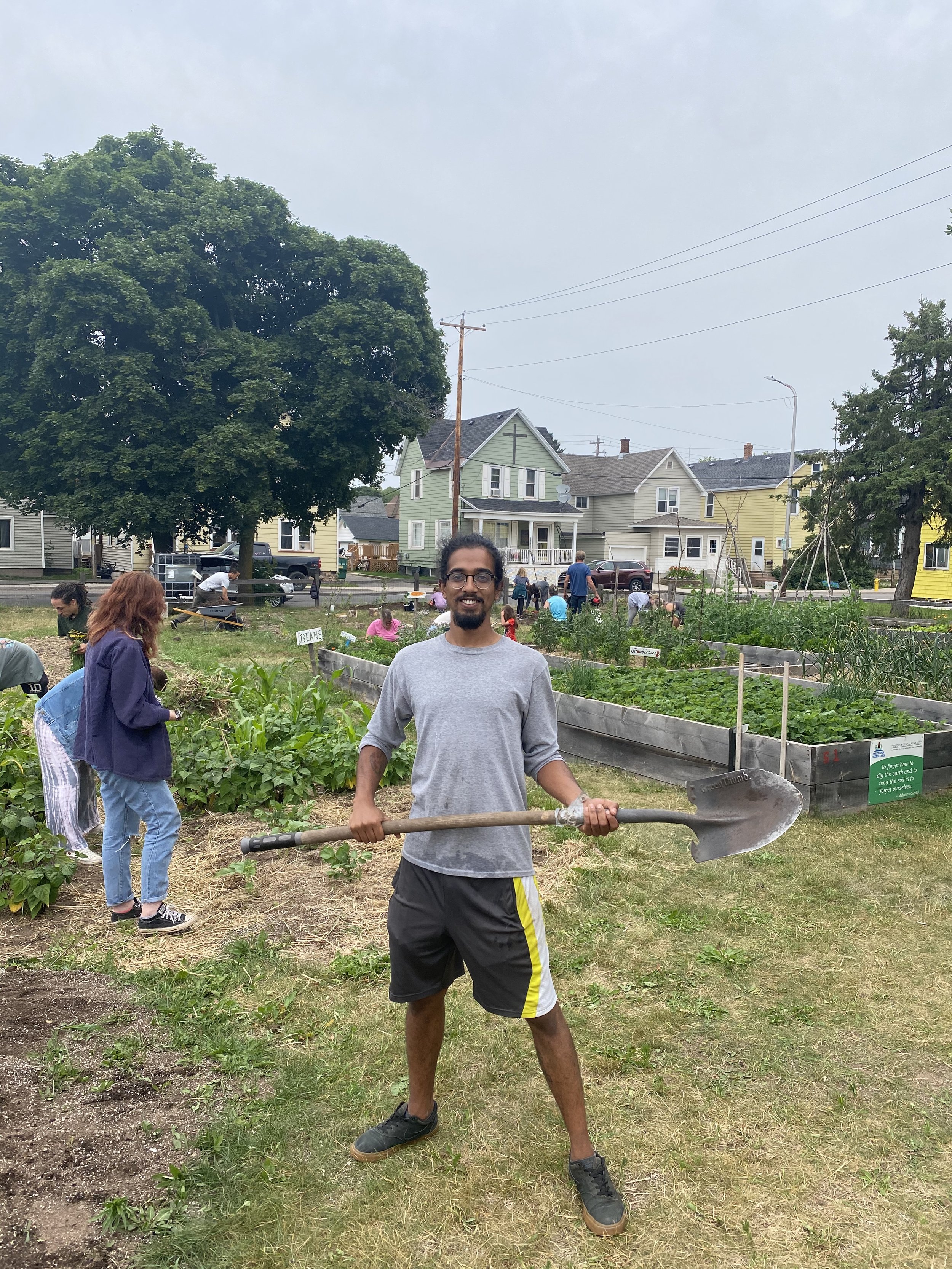 A man with glasses and a gray shirt holds a shovel in a community garden, with other people working in the garden and houses in the background.