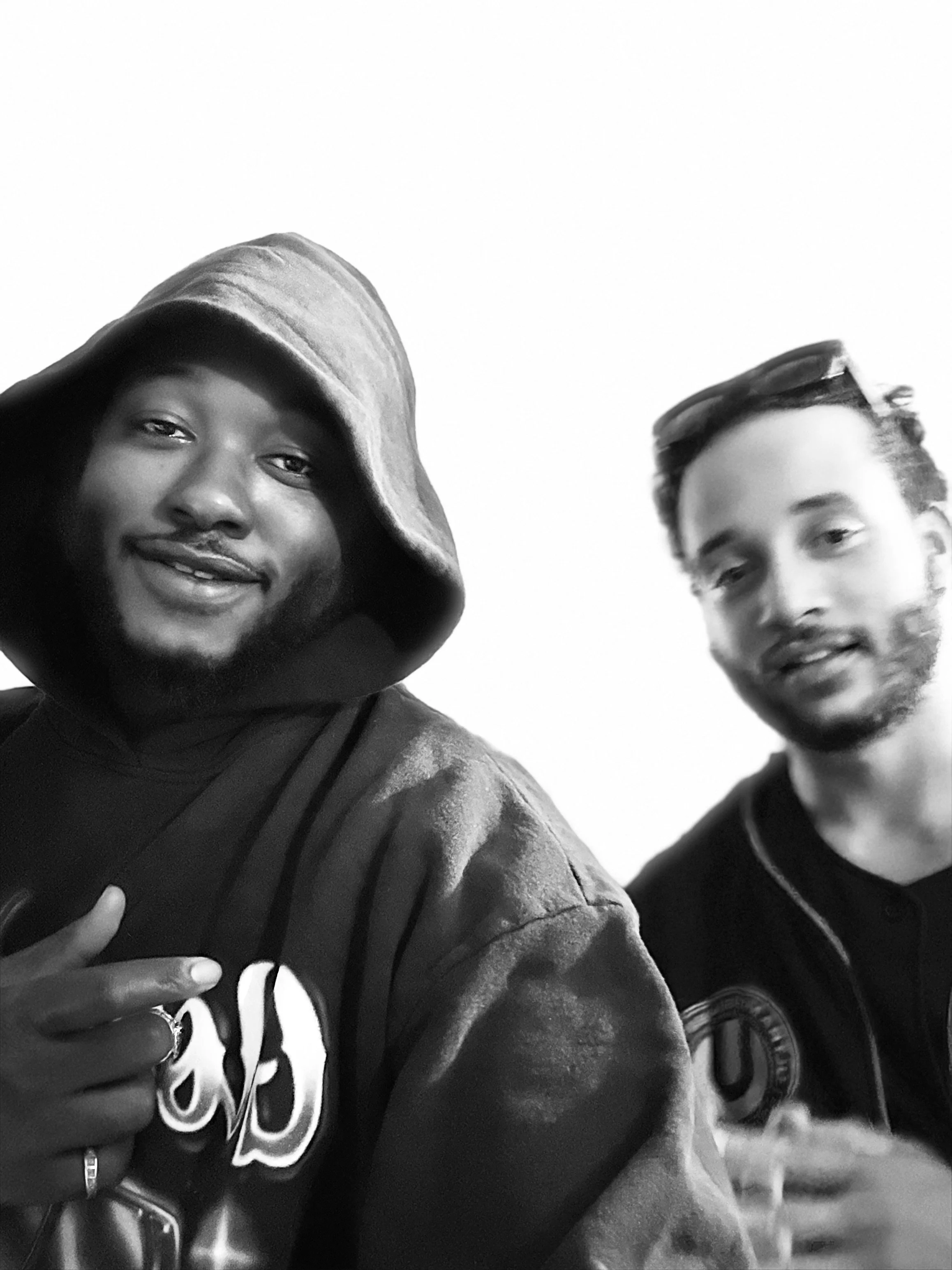 Two young people taking a selfie outdoors in black and white. The person on the left is a woman wearing a hoodie and making a peace sign. The person on the right is a man wearing sunglasses on his head and a dark sweatshirt.