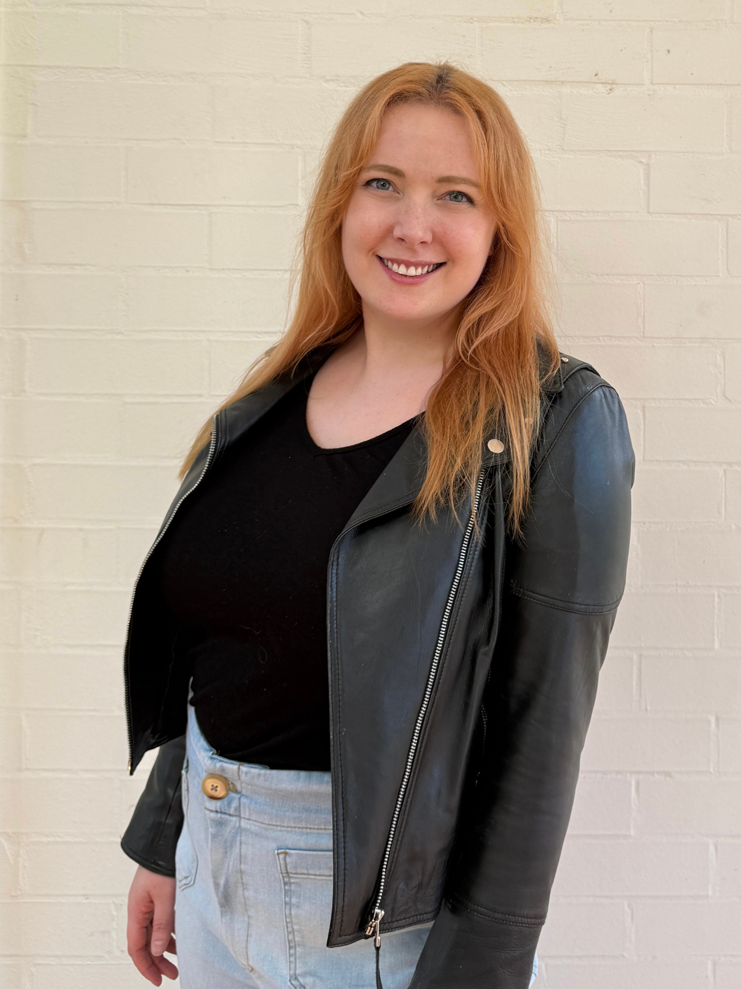 Jackie Money, marketing and publishing consultant, smiling in a black leather jacket and jeans against a white brick wall.