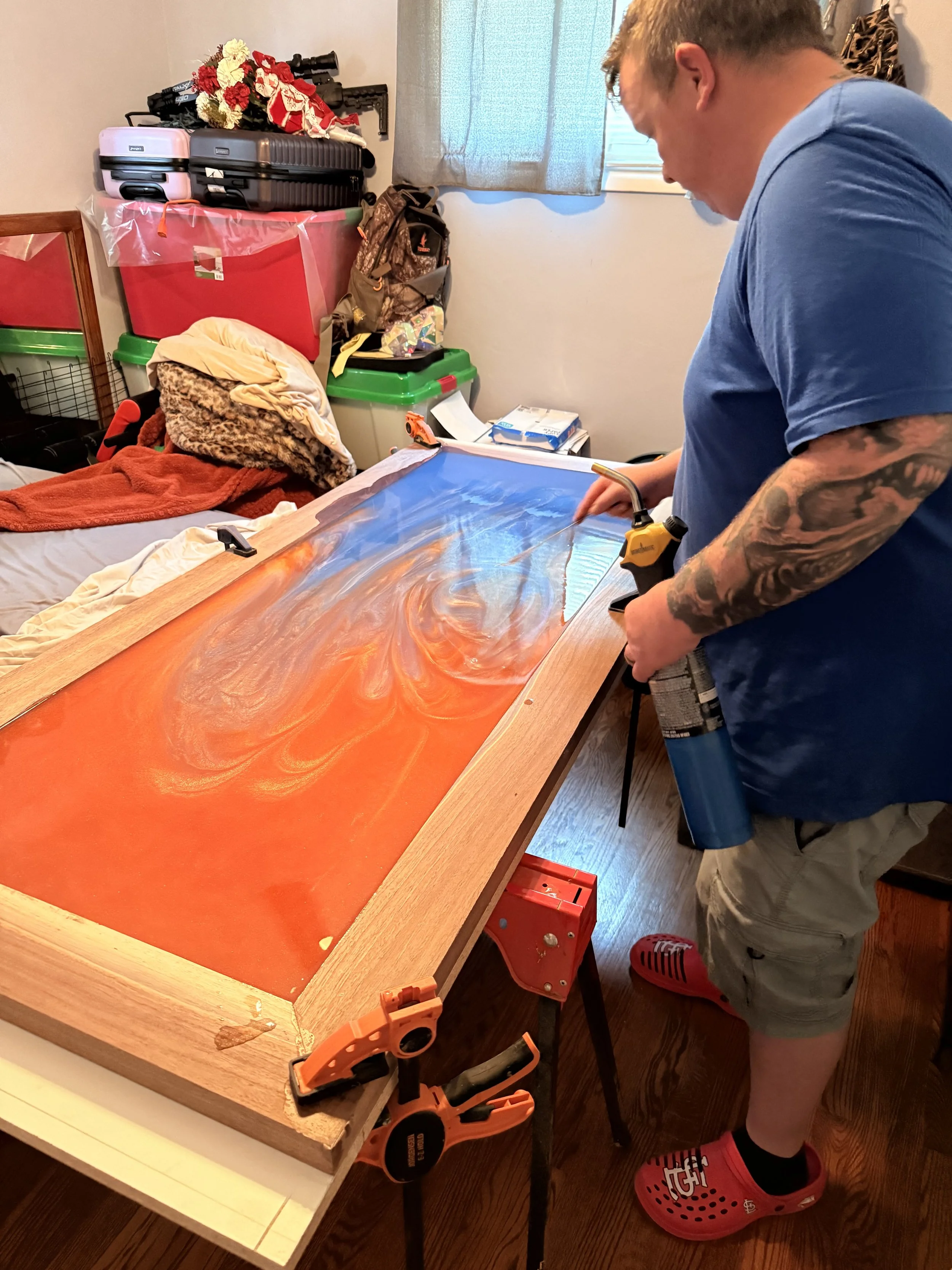 A person working on a furniture project, attaching a glass top to a wooden table. The person is wearing a blue shirt, shorts, and red Crocs, and has tattoos on their arms. The workspace has a bed or couch with blankets and pillows, storage containers