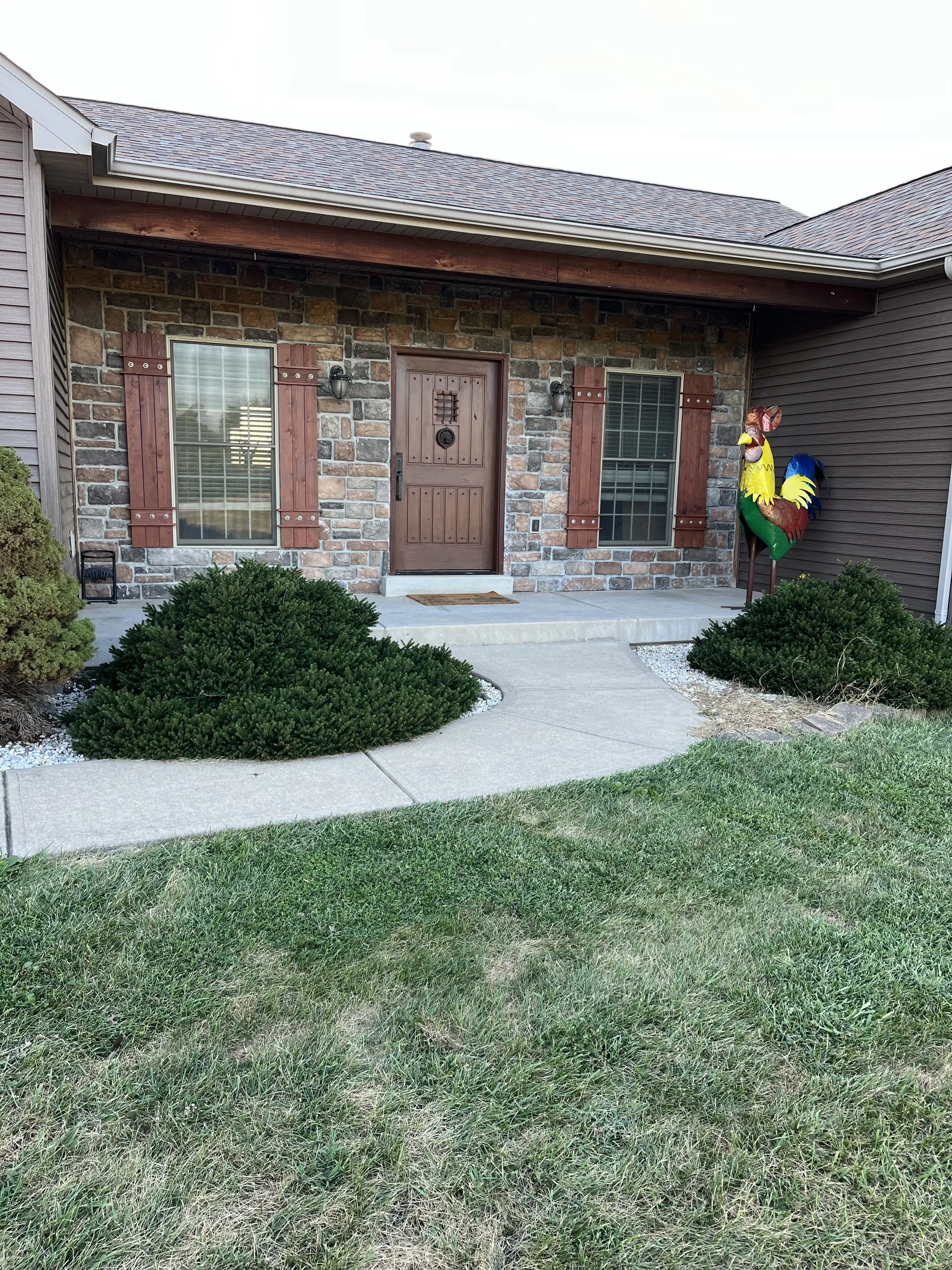 Front porch of a house with a brick facade, wooden shutters, and a decorative rooster sculpture on the right side. Green shrubs and a grassy lawn are in the foreground.