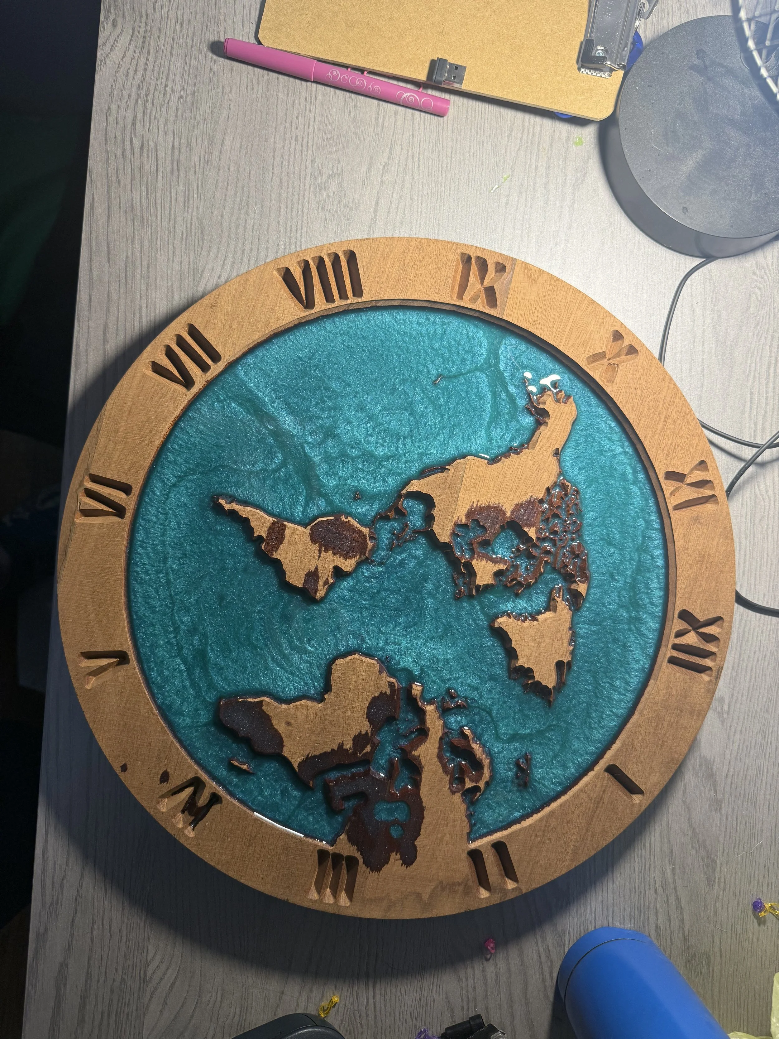 A decorative wall clock with a wooden frame featuring Roman numerals. The clock's face is designed with a map of the world in turquoise, with landmasses in a darker tone. The clock is placed on a gray wooden surface, surrounded by various office supp