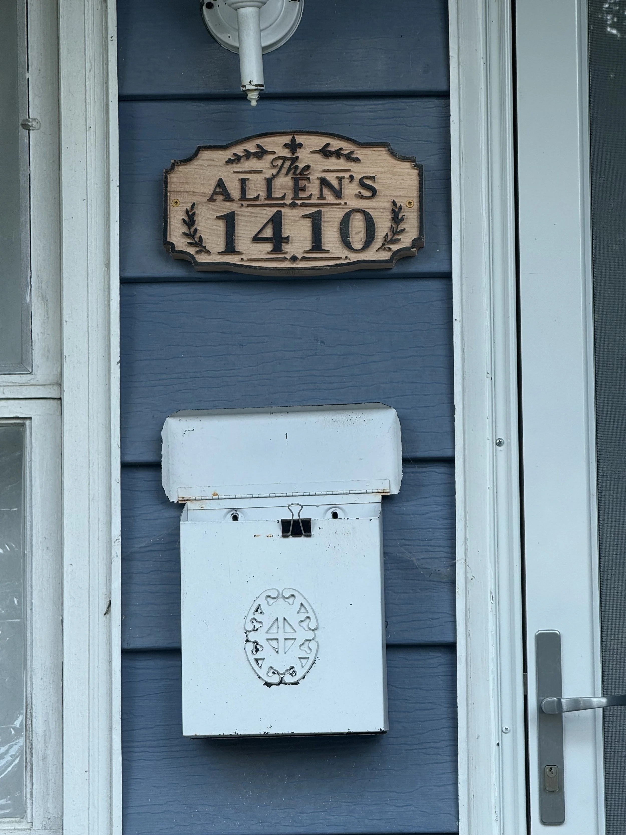 A house front door with a blue exterior wall, a metal mailbox, a wooden sign displaying the house number 1410 and the family name Allens, and a doorbell to the right.