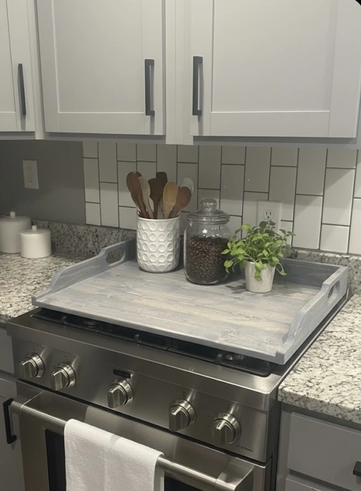 Kitchen stove with a tray holding a potted plant, jar of coffee beans, and a container with kitchen utensils, white cabinets above, granite countertop.