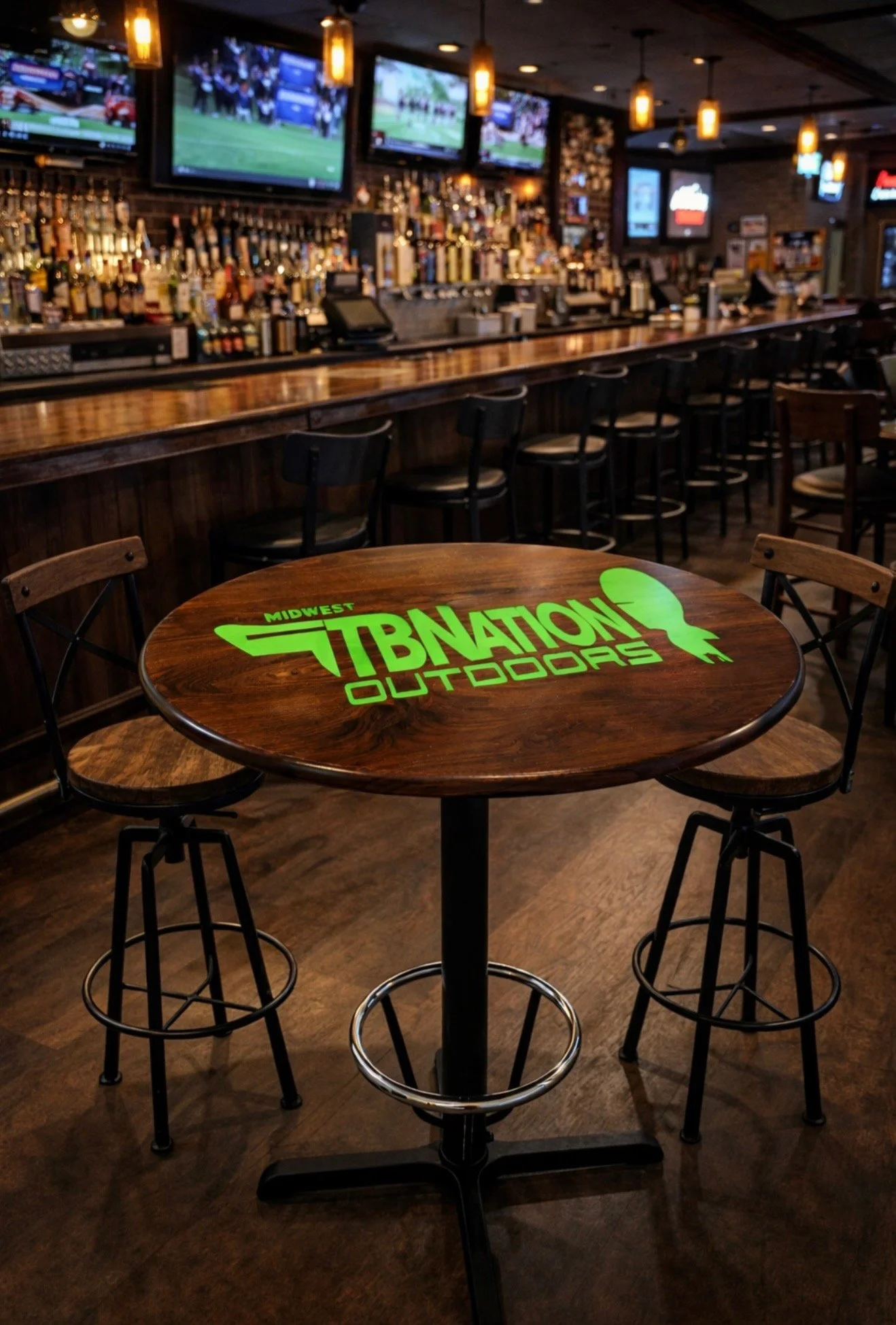 A wooden table with a bright green logo of 'Midwest TBNATION Outdoors' in a bar or restaurant. Bar stools are around the table. The background shows a bar counter with numerous bottles and multiple TV screens mounted on the wall displaying sports.