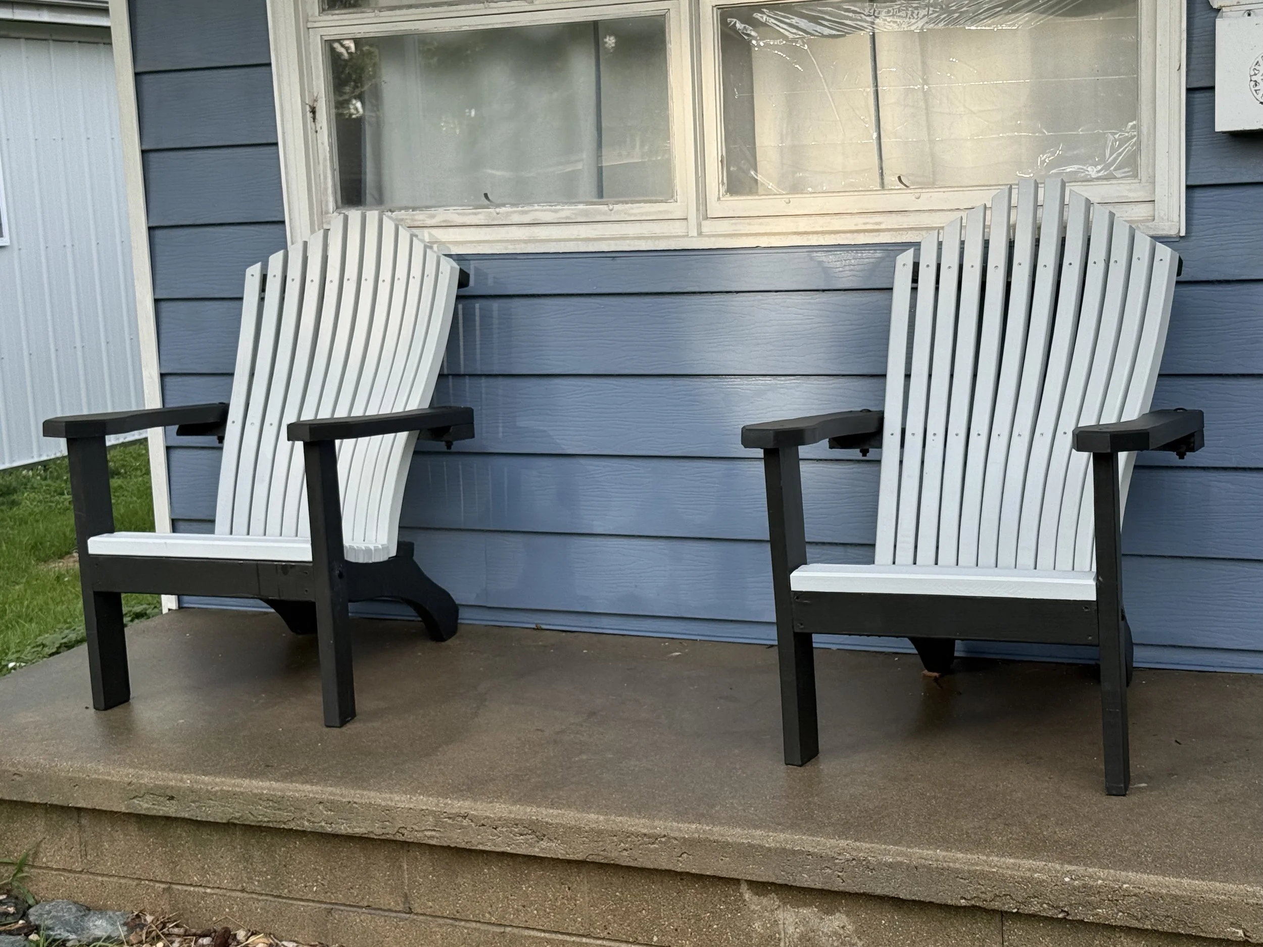 Two Adirondack chairs on a porch in front of a blue house with a window and siding.