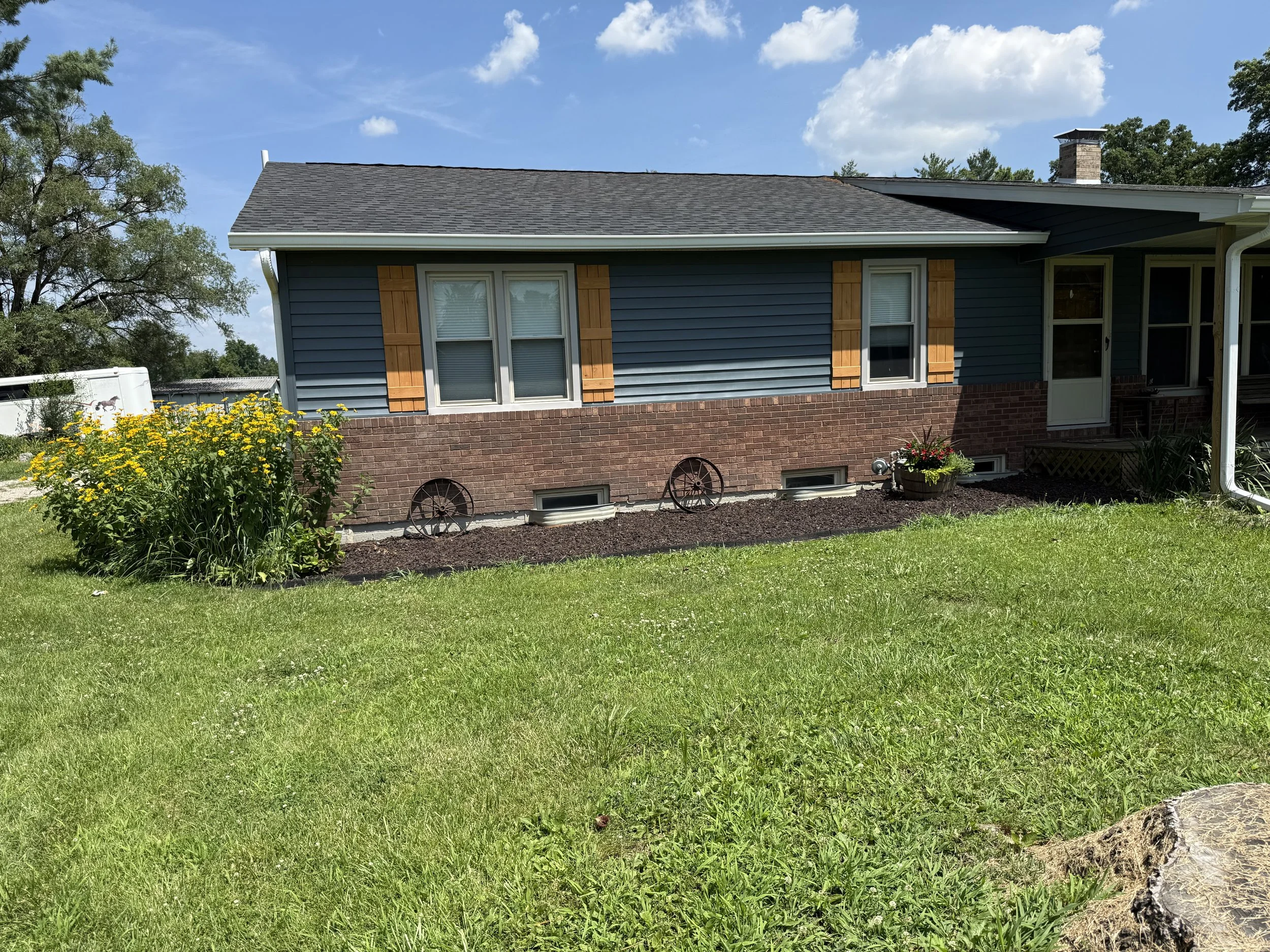 A house with blue siding, wooden shutters, and a brick foundation, with a garden in front, clear blue sky with a few clouds.