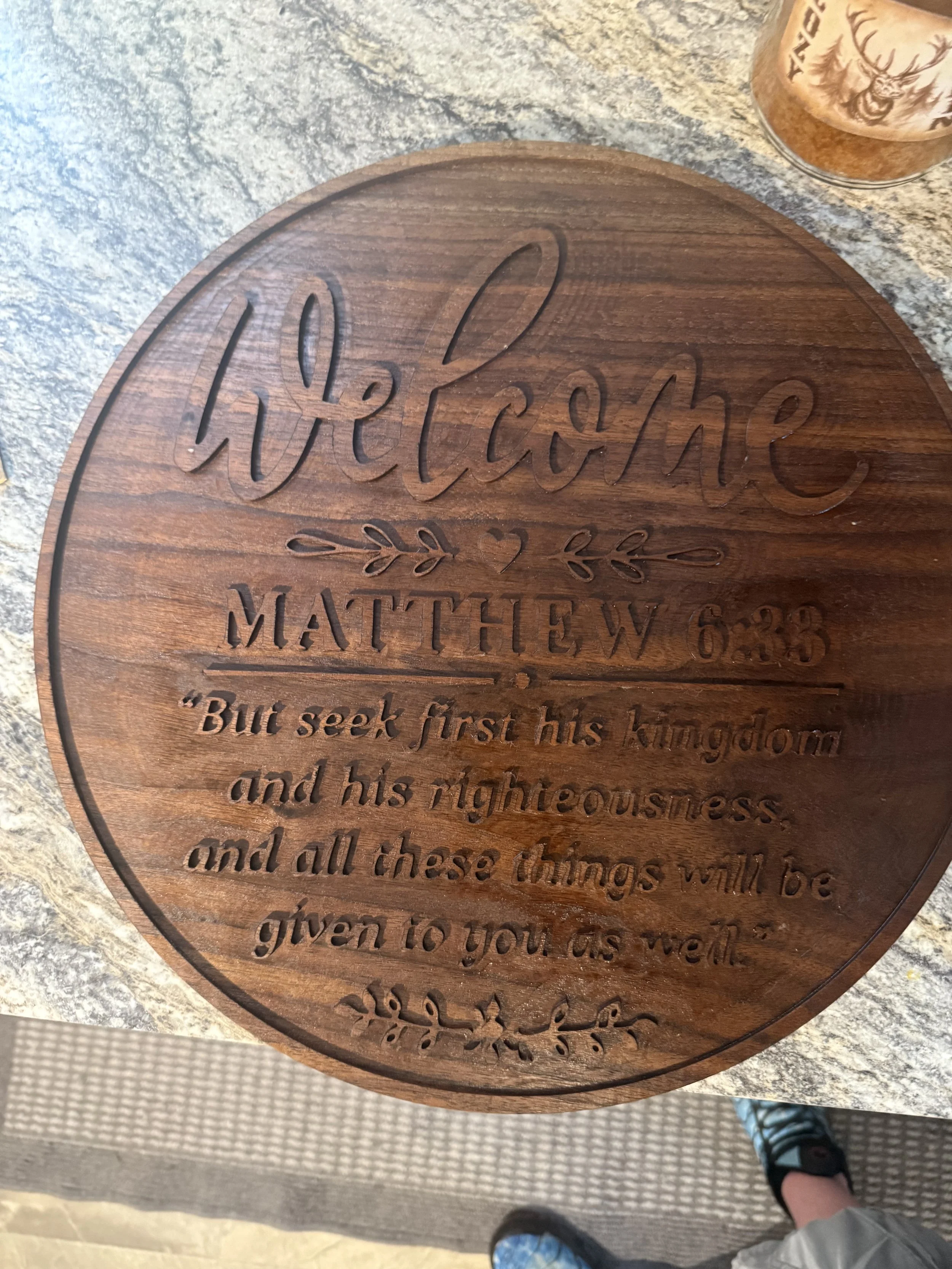 A round wooden plaque with the Bible verse Matthew 6:33 engraved on it. Part of a person's leg, foot in a sandal, is visible at the bottom of the image. The plaque is on a textured surface, likely a table or countertop, with a container holding a sna