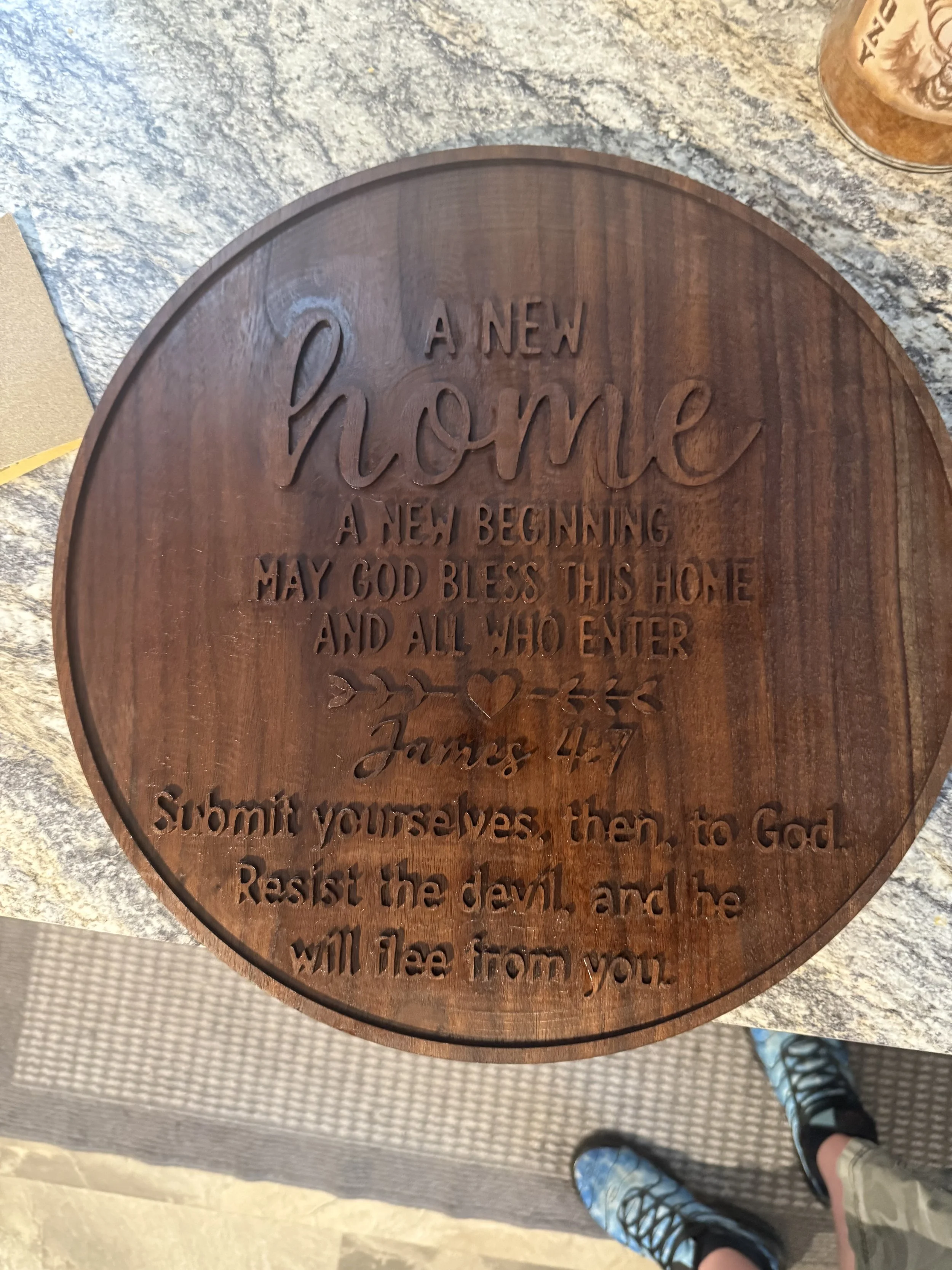 Wooden plaque with engraved text reading 'A new home, a new beginning. May God bless this home and all who enter. James 4:7'. Part of a person's feet in sneakers and shorts are visible, standing on a carpeted and tiled floor.