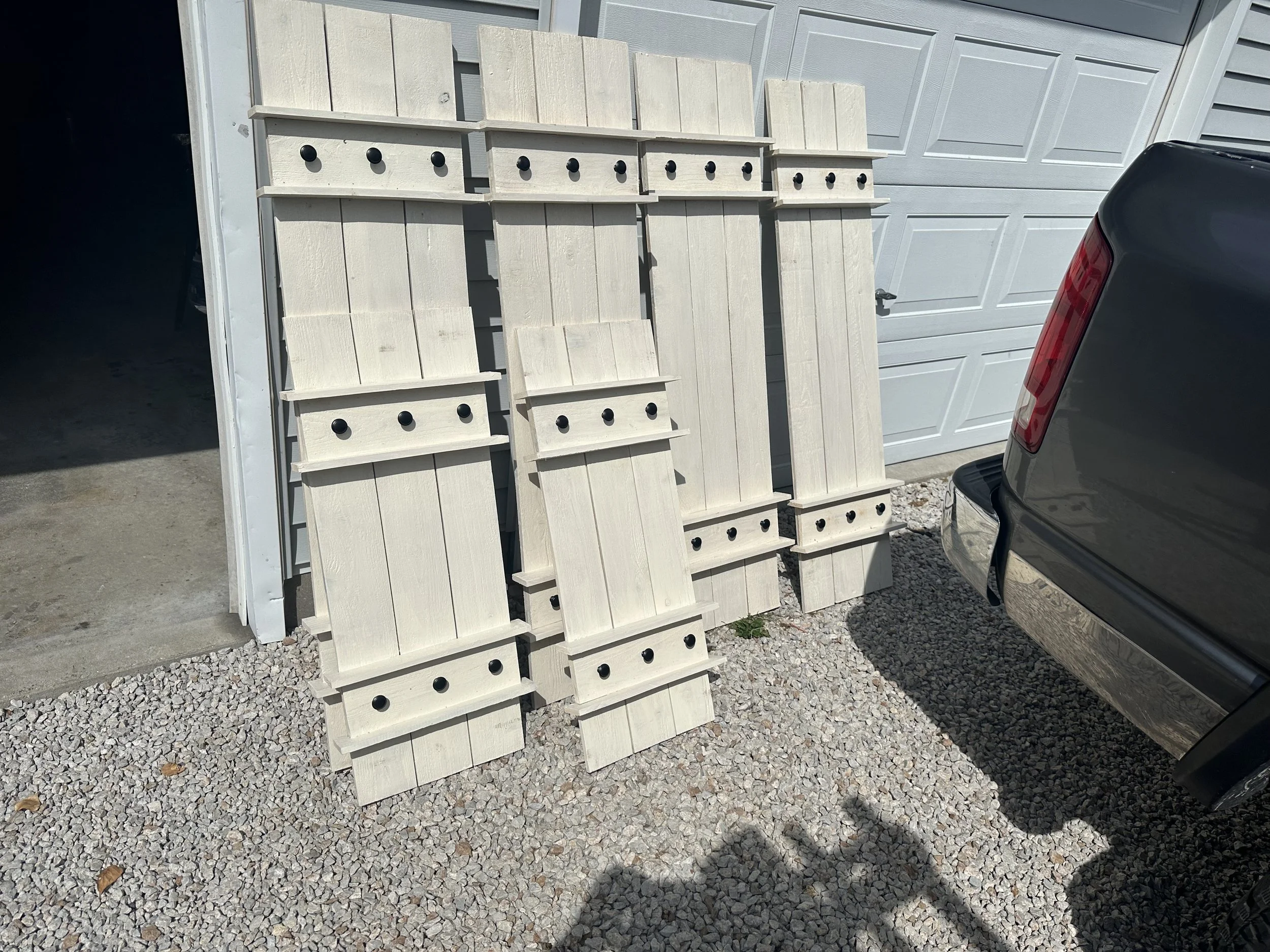 A display of white wooden furniture pieces with black knobs, leaning against a garage door on a gravel driveway, with part of a black vehicle visible in the foreground.