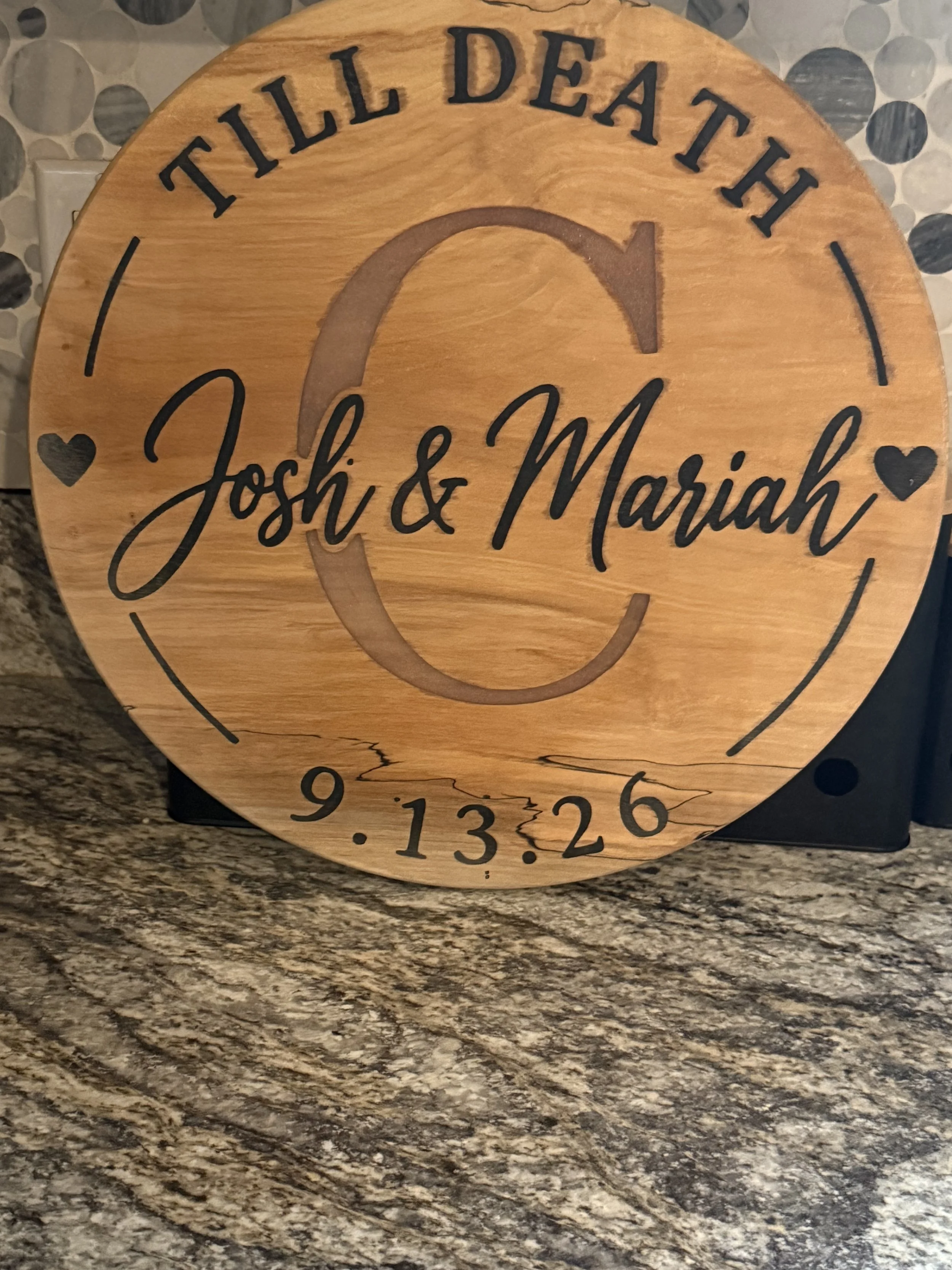 A wooden clock with black markings and numbers, including a handwritten message 'Till Death' at the top and the names 'Josh & Mariah' in the center, with a heart symbol beside each name, placed on a granite countertop.