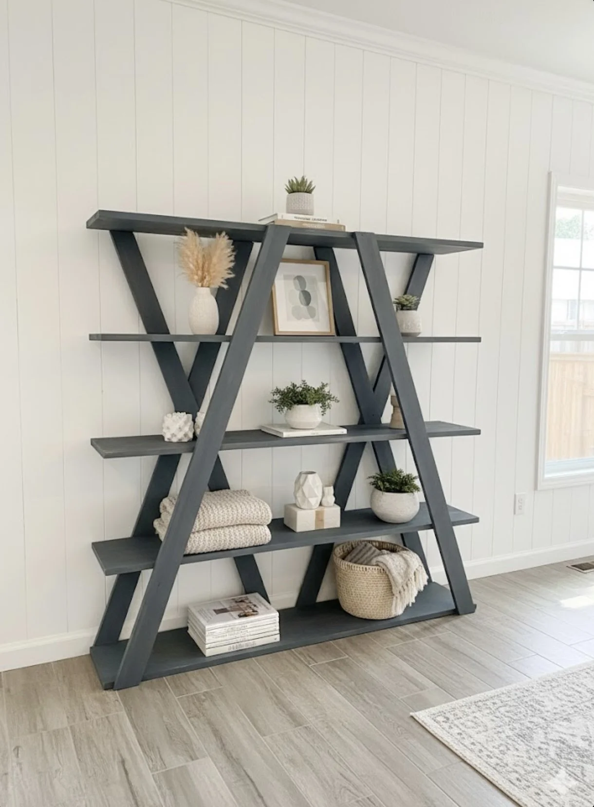 Gray A-frame bookshelf decorated with plants, books, and decorative objects, positioned against a white wall near a window in a bright living space.