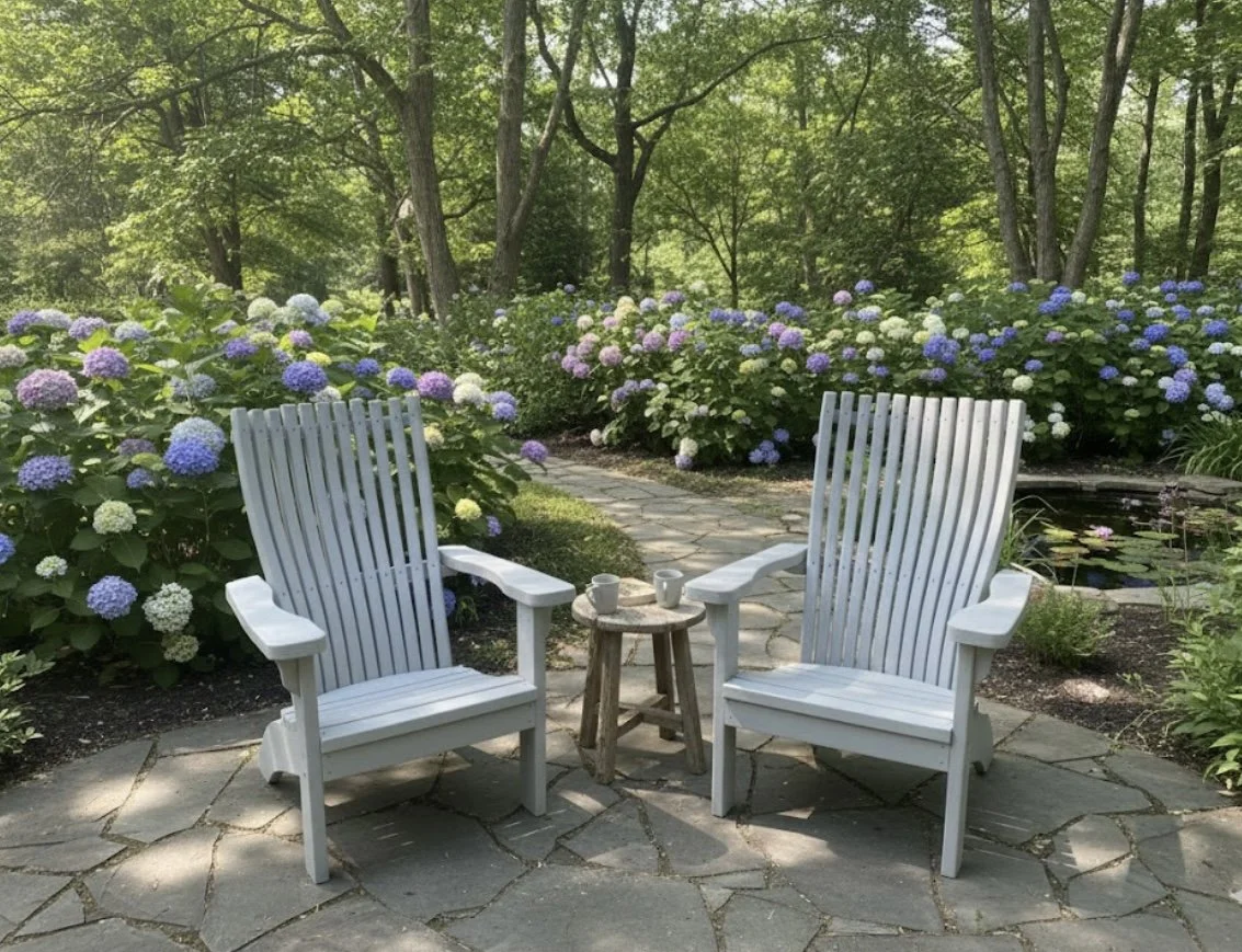 Two white Adirondack chairs face each other with a small wooden table in between, set on a stone patio surrounded by colorful hydrangea bushes in a lush green garden.